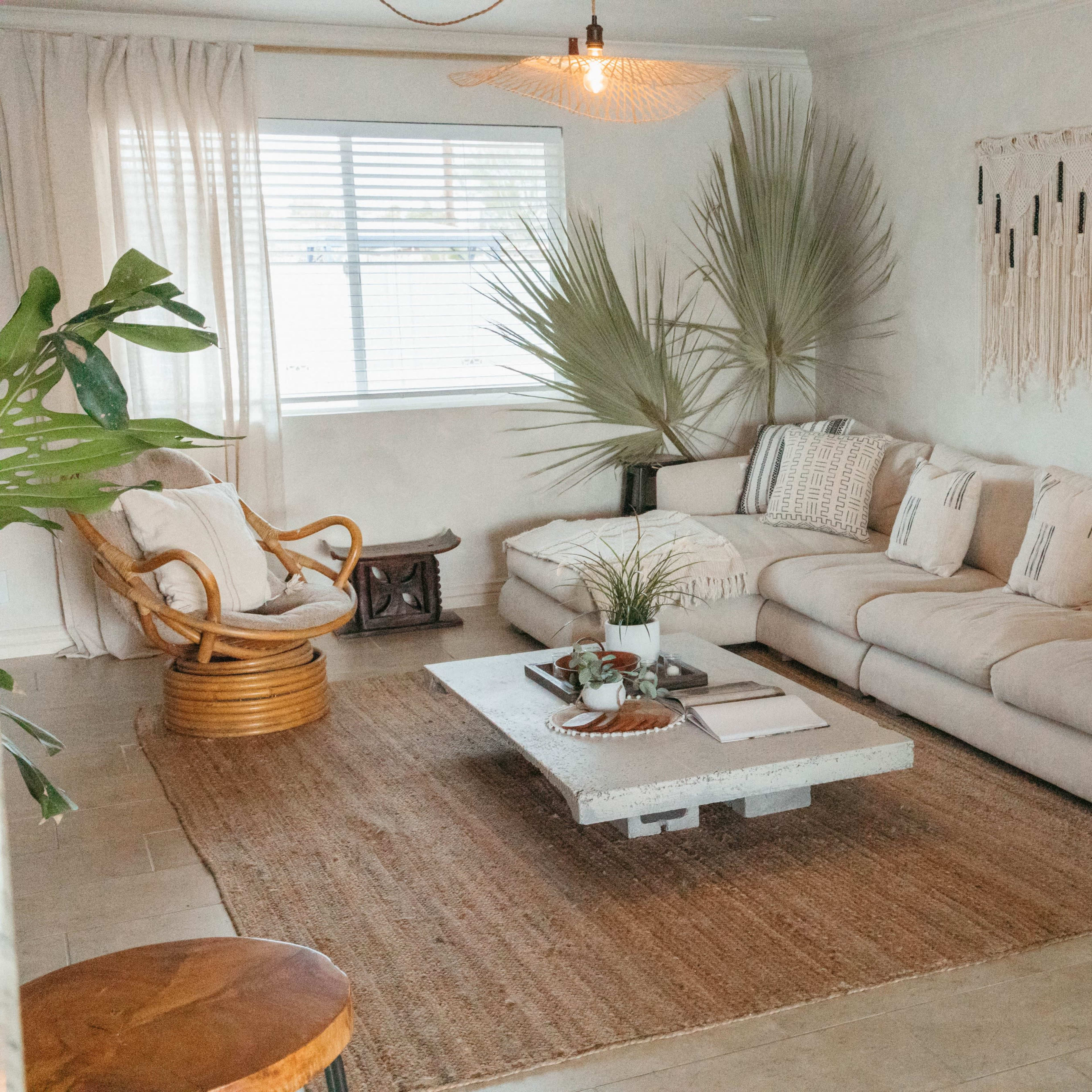A cozy living room featuring a large light-colored sofa, a low coffee table, and various potted plants, with a woven rug on the floor and natural light coming through a window.