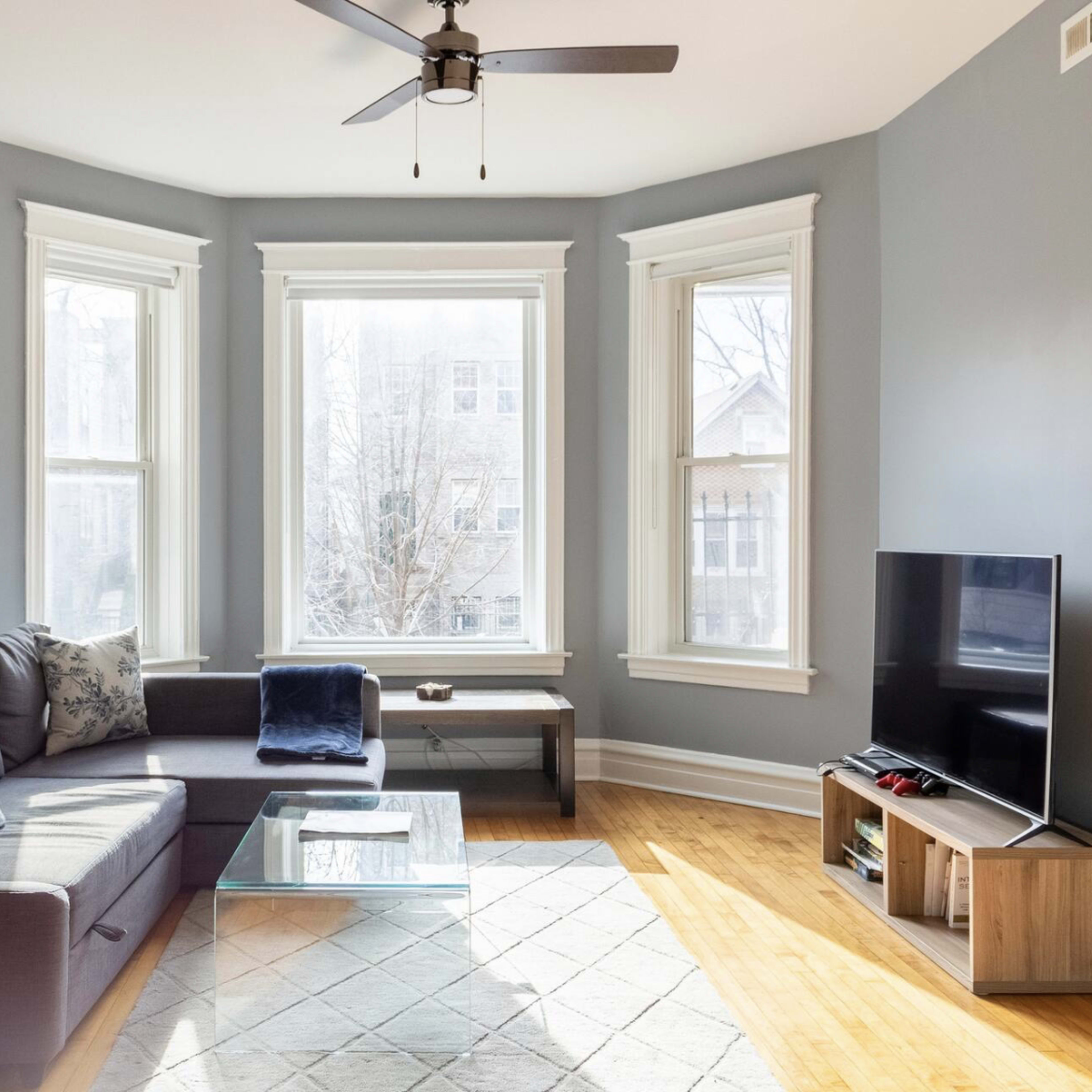 A living room with a sectional sofa, a coffee table, large windows allowing natural light, and a television on a stand against a wall.