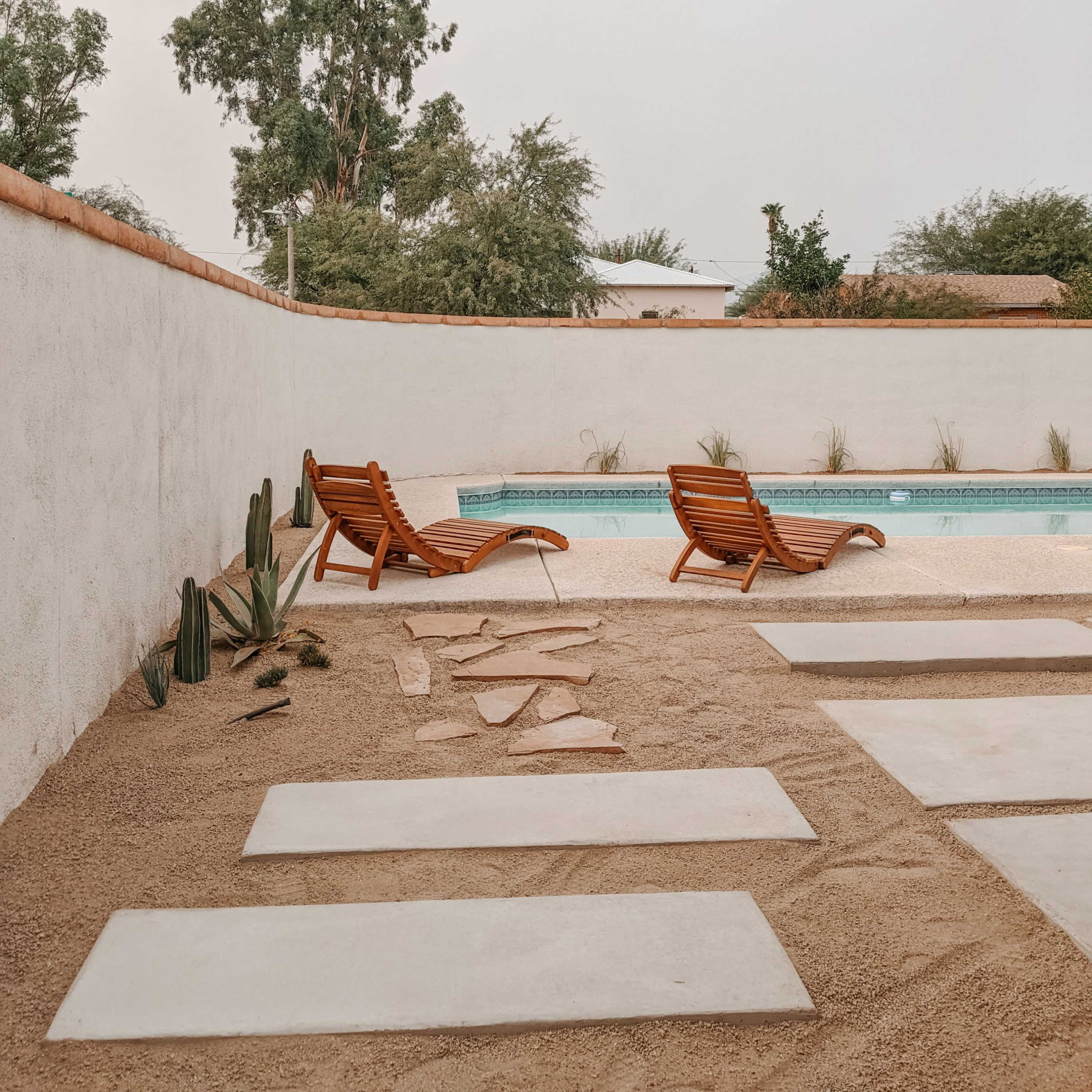 A pool area with two wooden lounge chairs, stone pavement, and cacti, surrounded by a white wall and greenery.