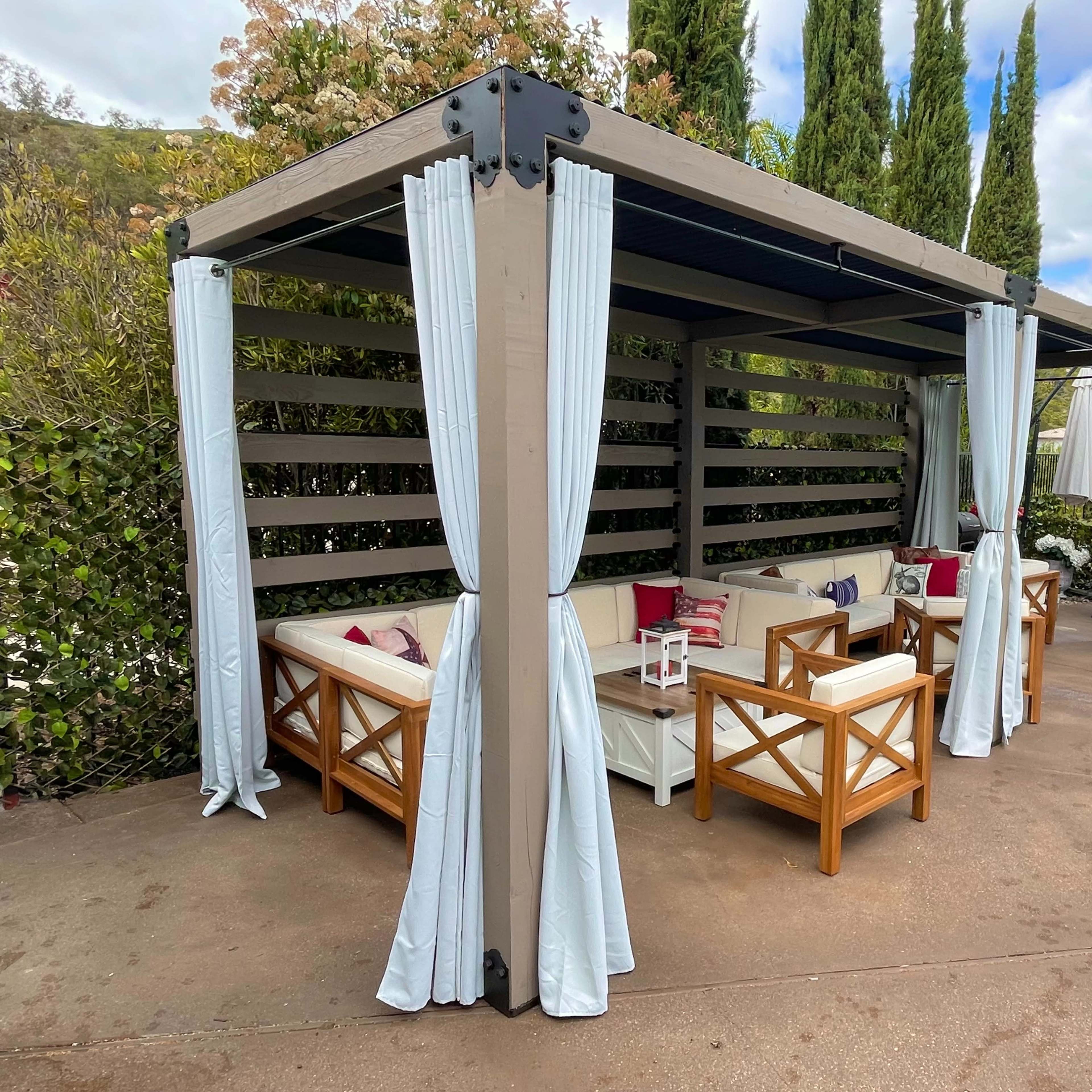 A shaded outdoor seating area is enclosed by curtains and features wooden furniture beside a swimming pool.