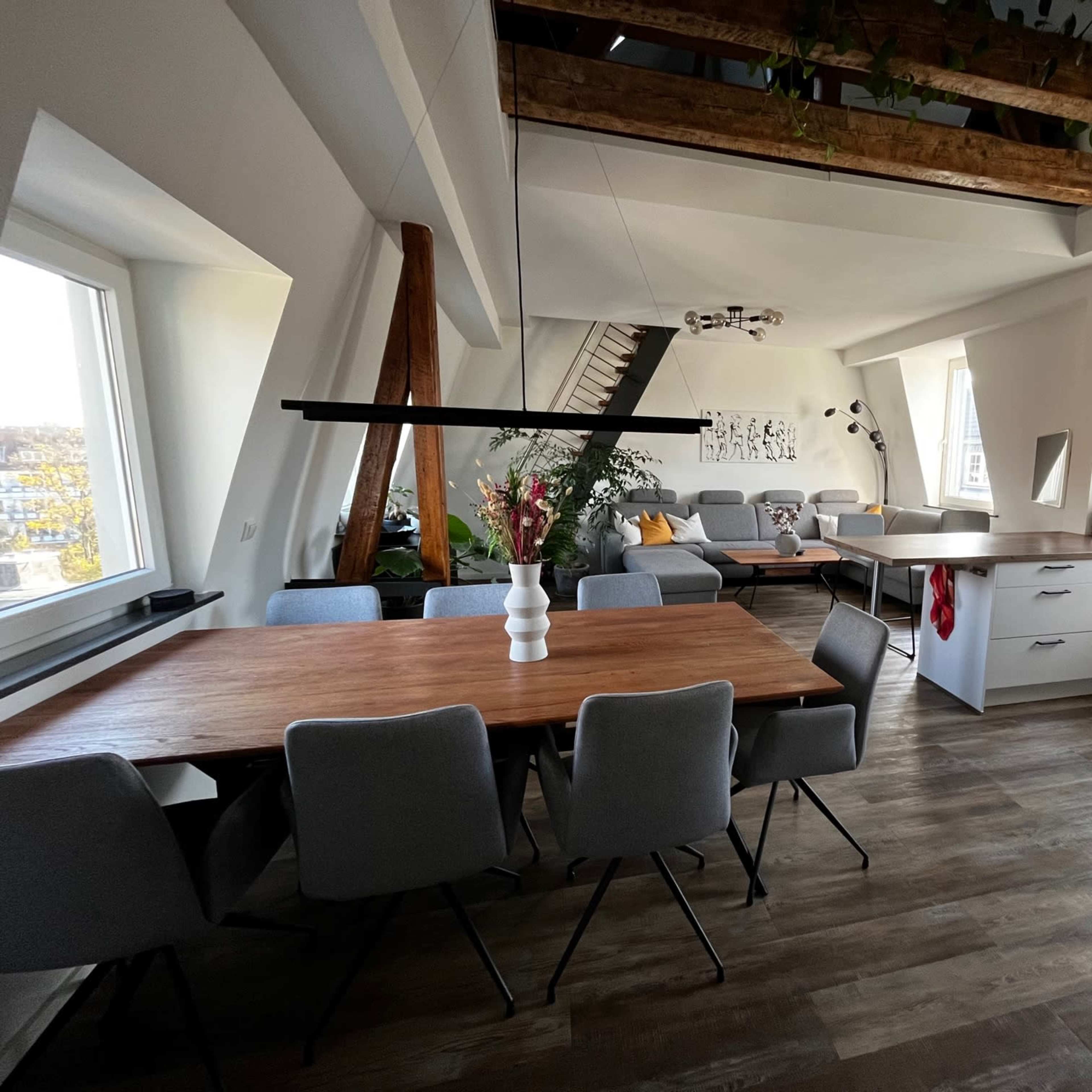 A modern kitchen and dining area featuring a wooden table with gray chairs, white cabinetry, and large windows providing natural light.
