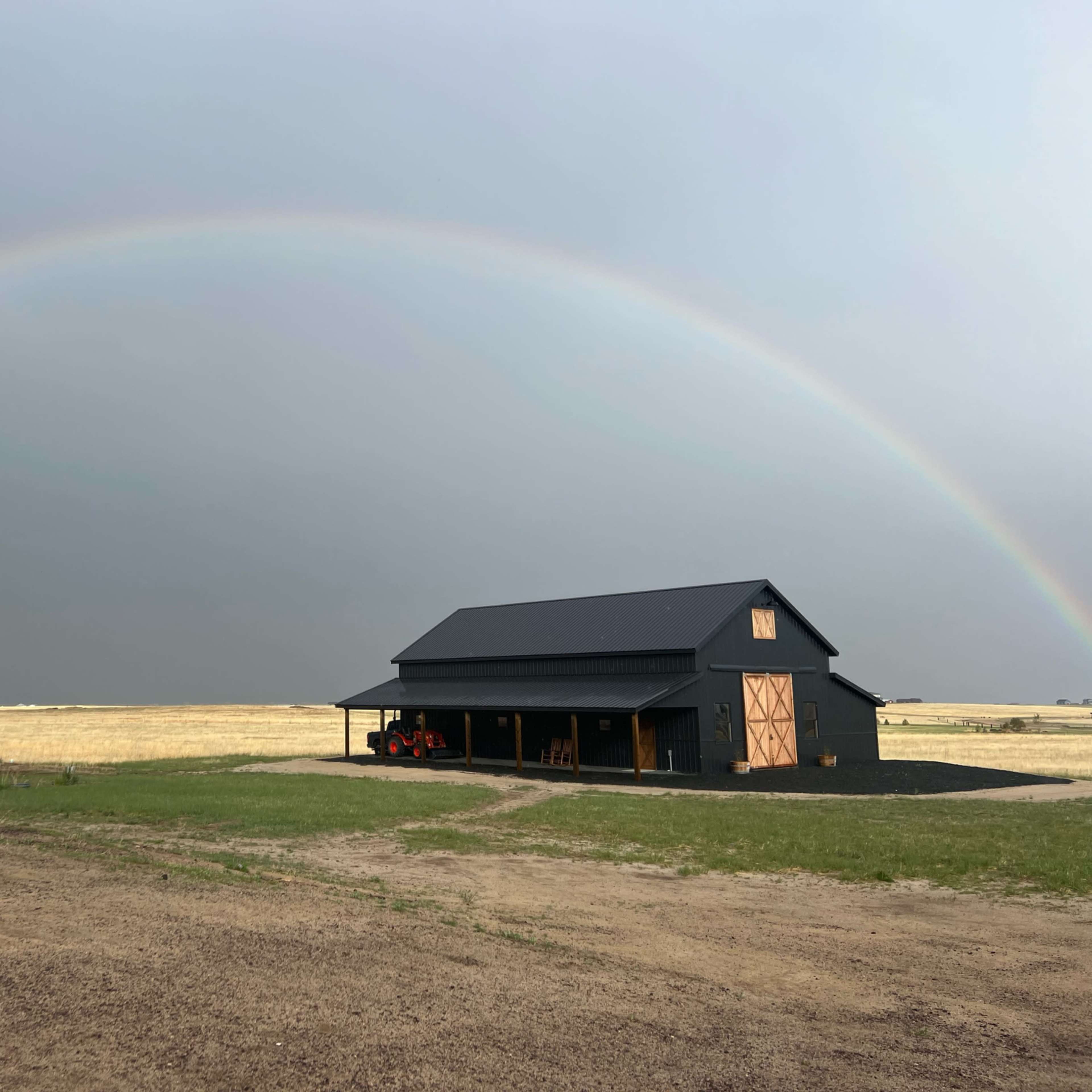 A black barn with a tractor parked nearby sits under a rainbow arching across a gray sky.