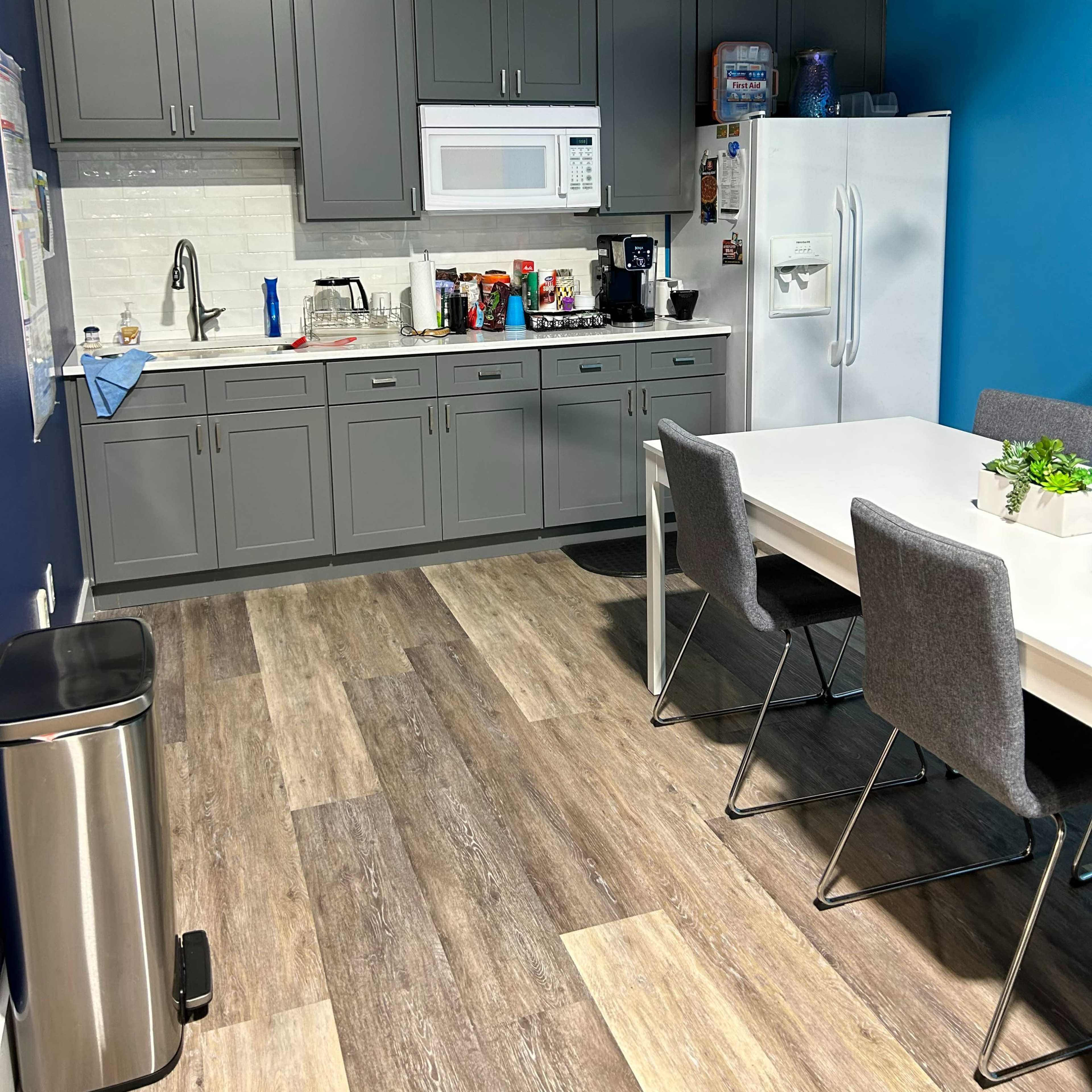 A modern kitchen area with gray cabinets, a white microwave and refrigerator, and a dining table with gray chairs on a wood-patterned floor.