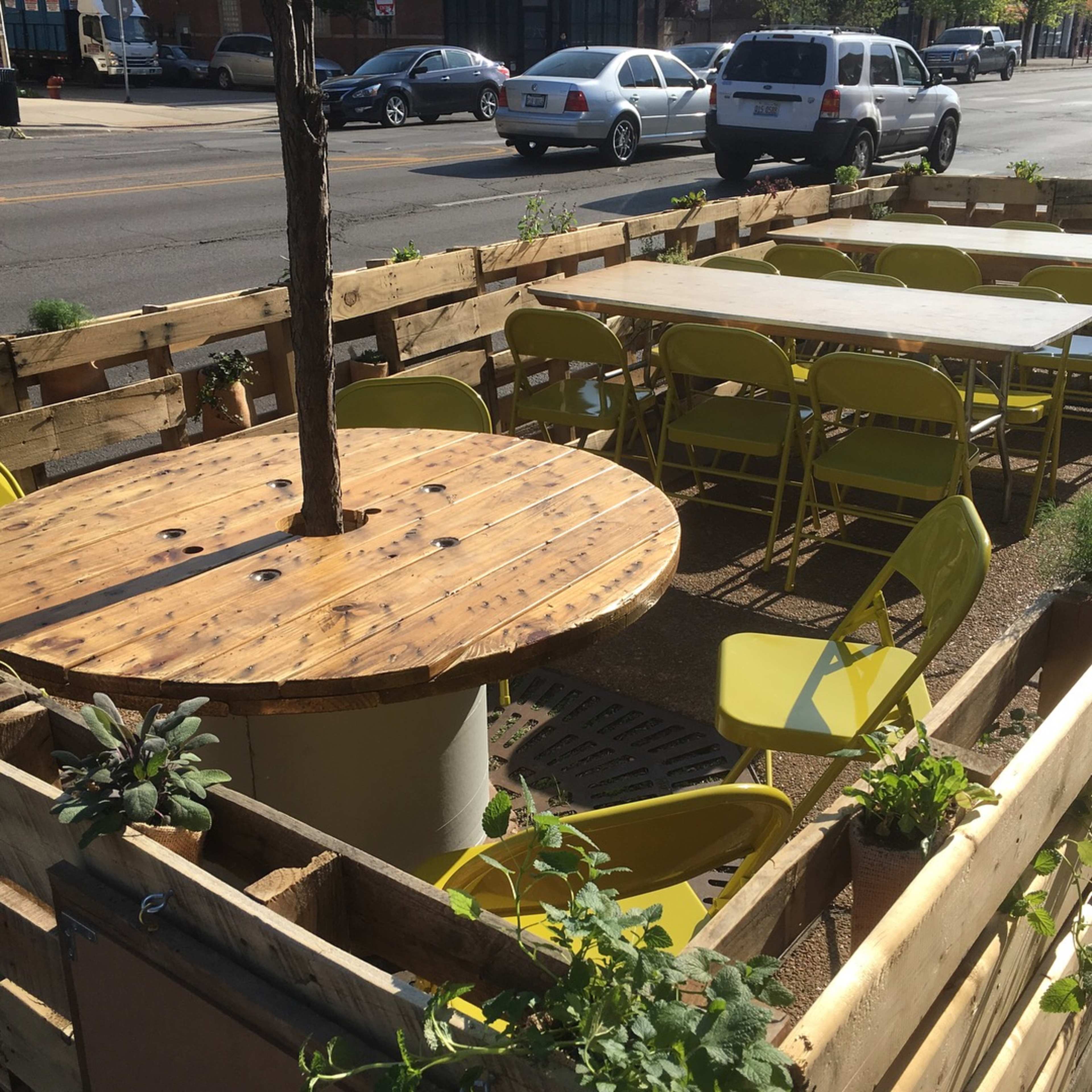 A wooden outdoor dining area with a round table featuring a tree at its center, surrounded by green folding chairs and a wooden enclosure.