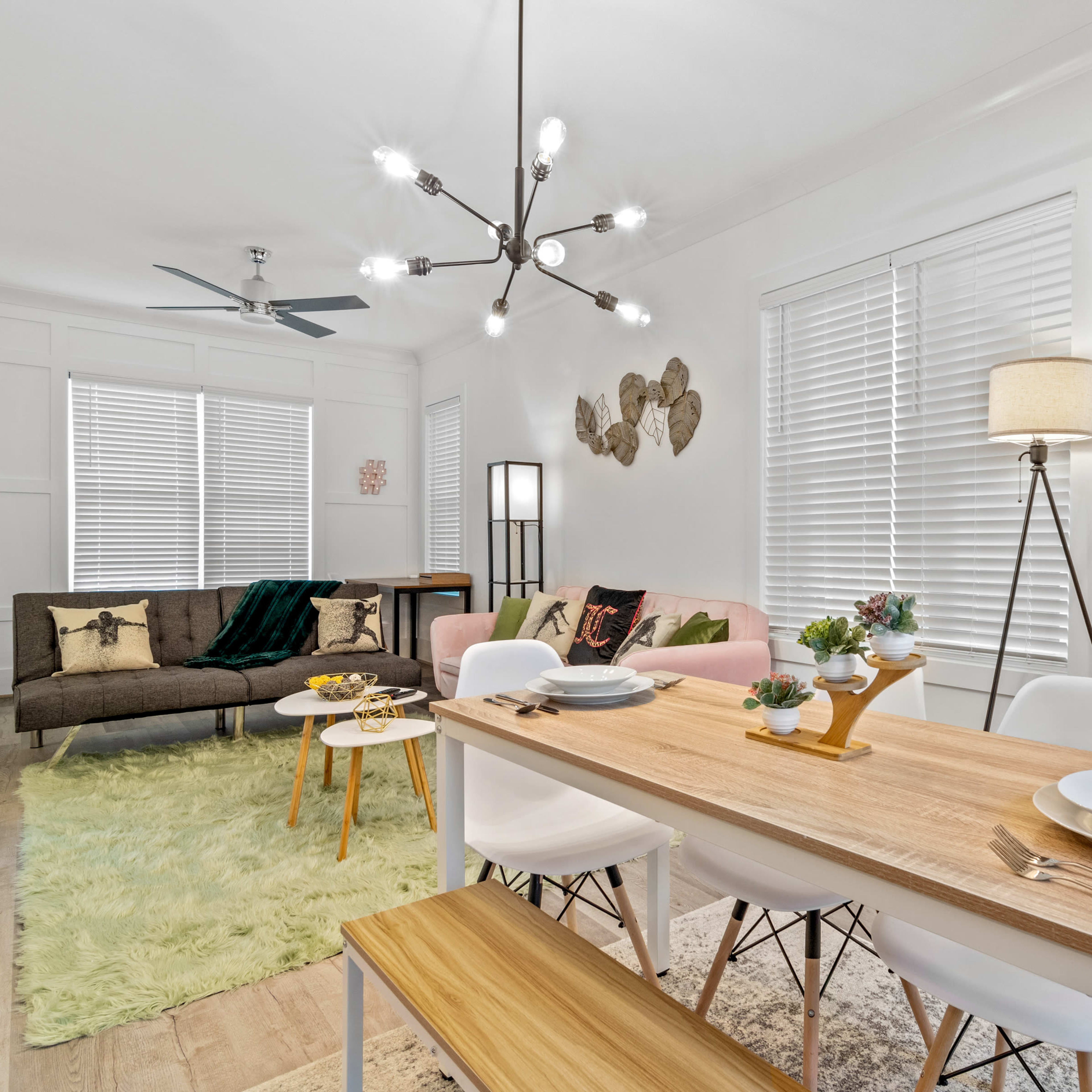 A modern living and dining area features a wooden table set for a meal, surrounded by white chairs, with a sofa, decorative items, and large windows in the background.