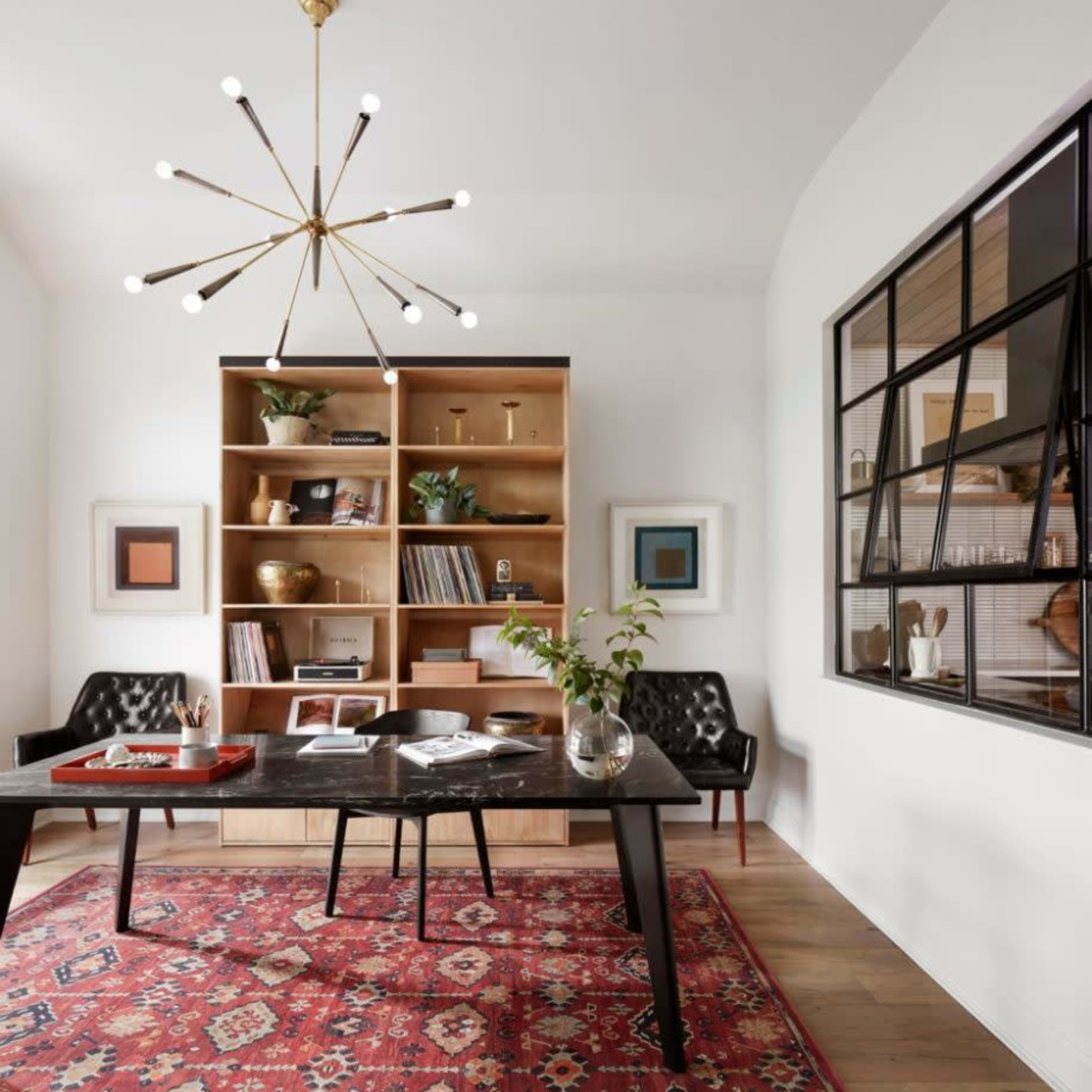A modern office features a large dark table, a bookshelf filled with books and decorative items, and a patterned rug on the hardwood floor.