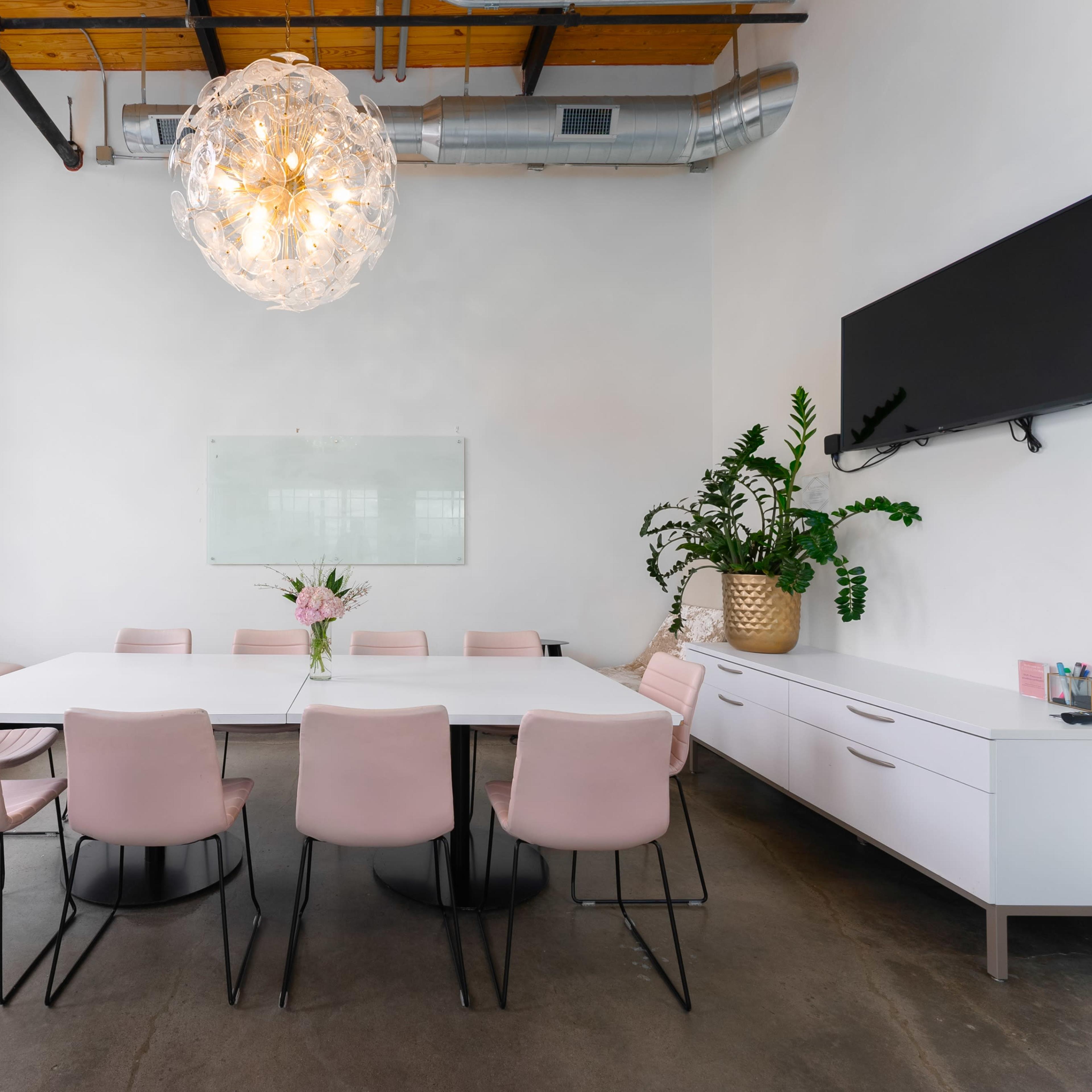 A modern meeting room features a large white table surrounded by pink chairs, illuminated by a decorative light fixture, with a plant and a whiteboard on the wall.