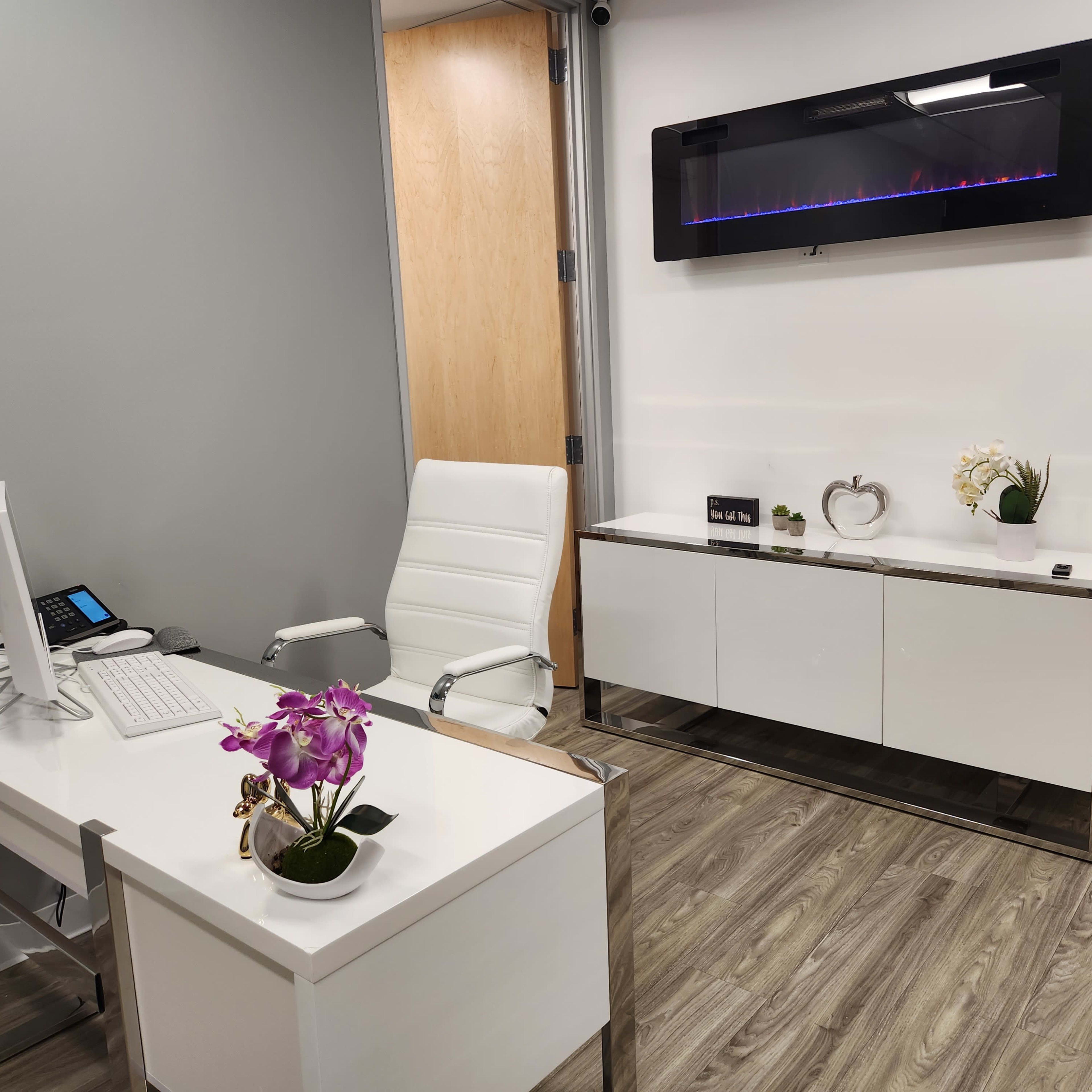 A modern office space with a white desk, an ergonomic chair, a sleek white cabinet, and a wall-mounted electric fireplace, all set against a light gray backdrop and wooden flooring.