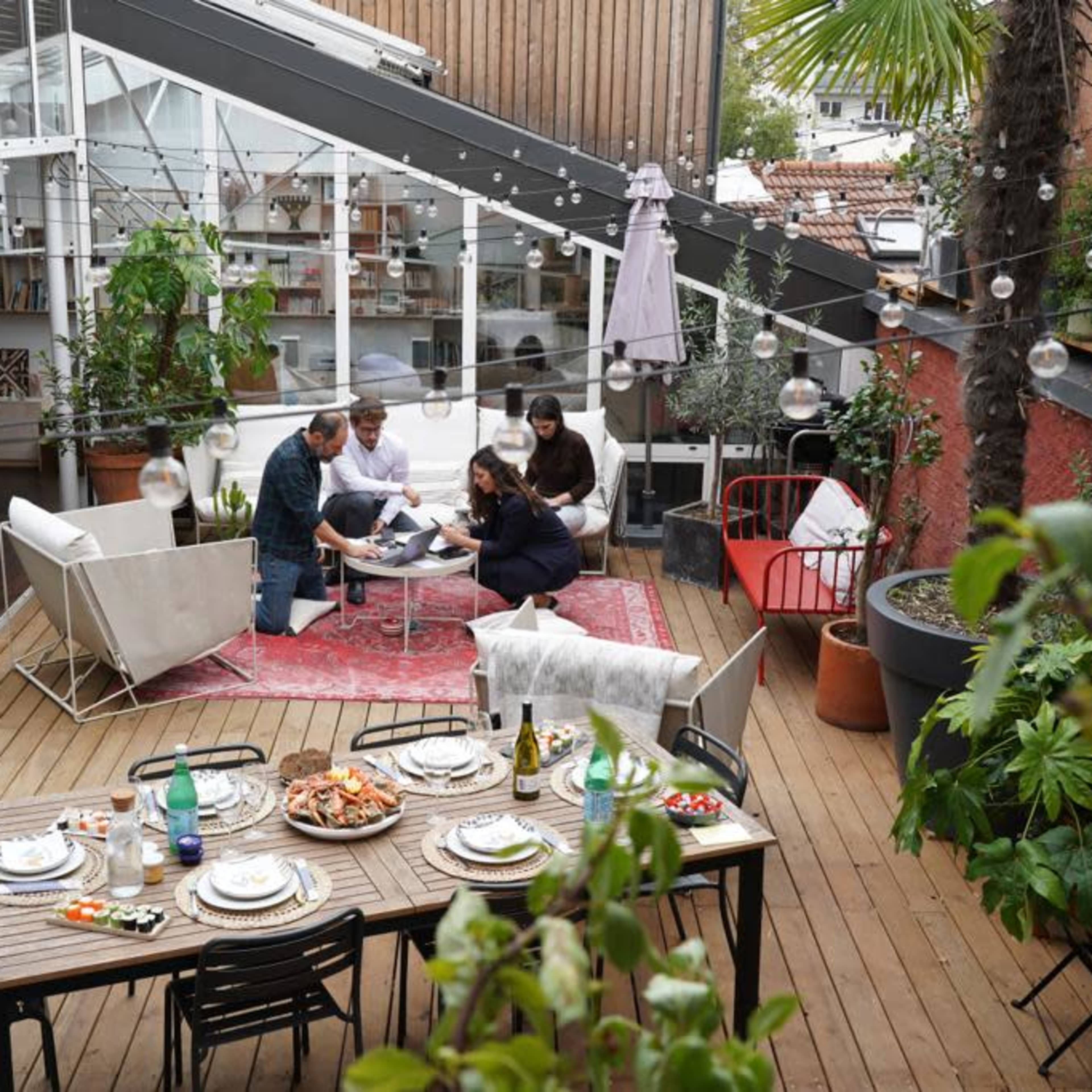 A group of five people gathers around a low table in an outdoor living space, surrounded by plants and string lights, with a dining table set for a meal nearby.