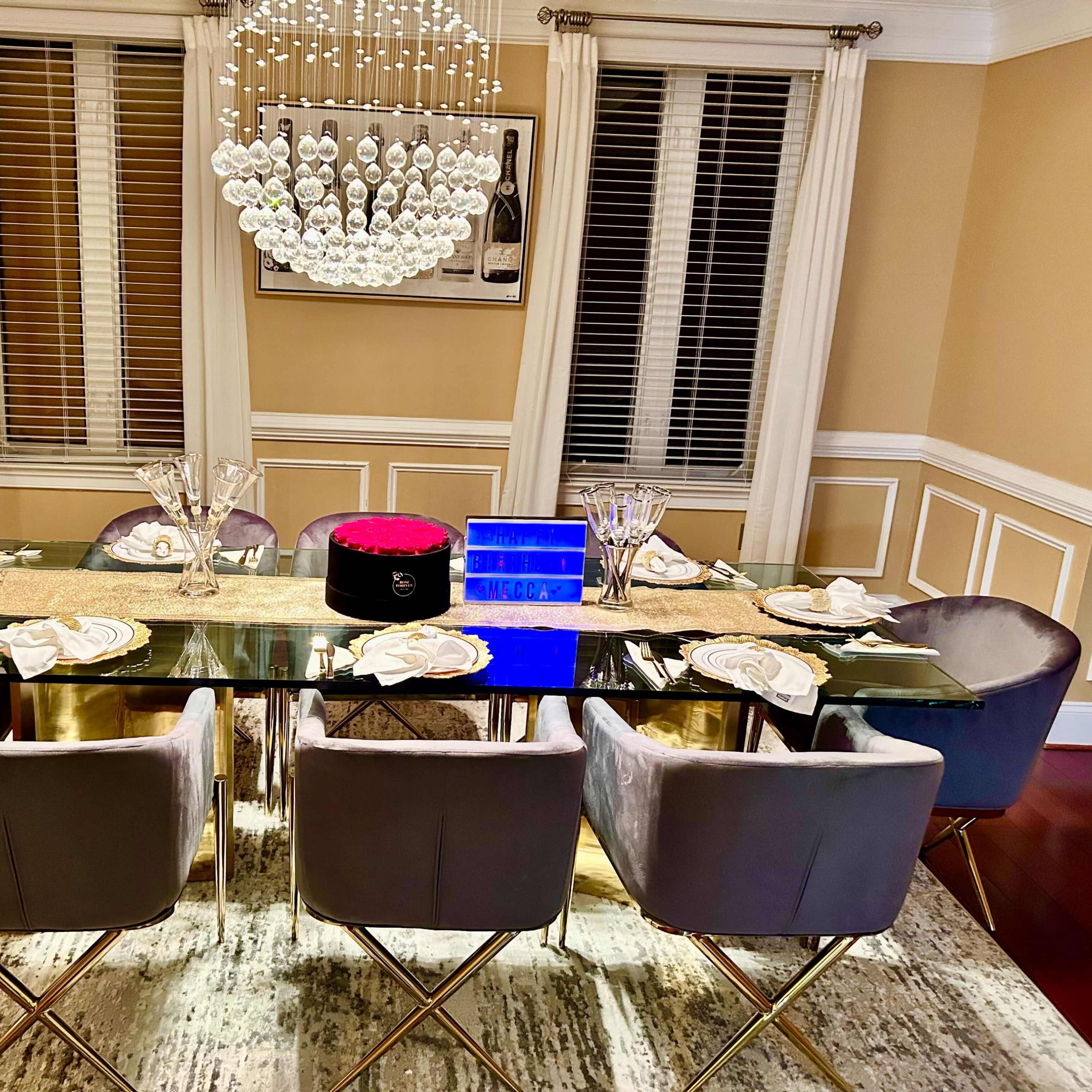 A glass dining table is set with elegant tableware and surrounded by upholstered chairs, beneath a modern chandelier in a well-decorated dining room.
