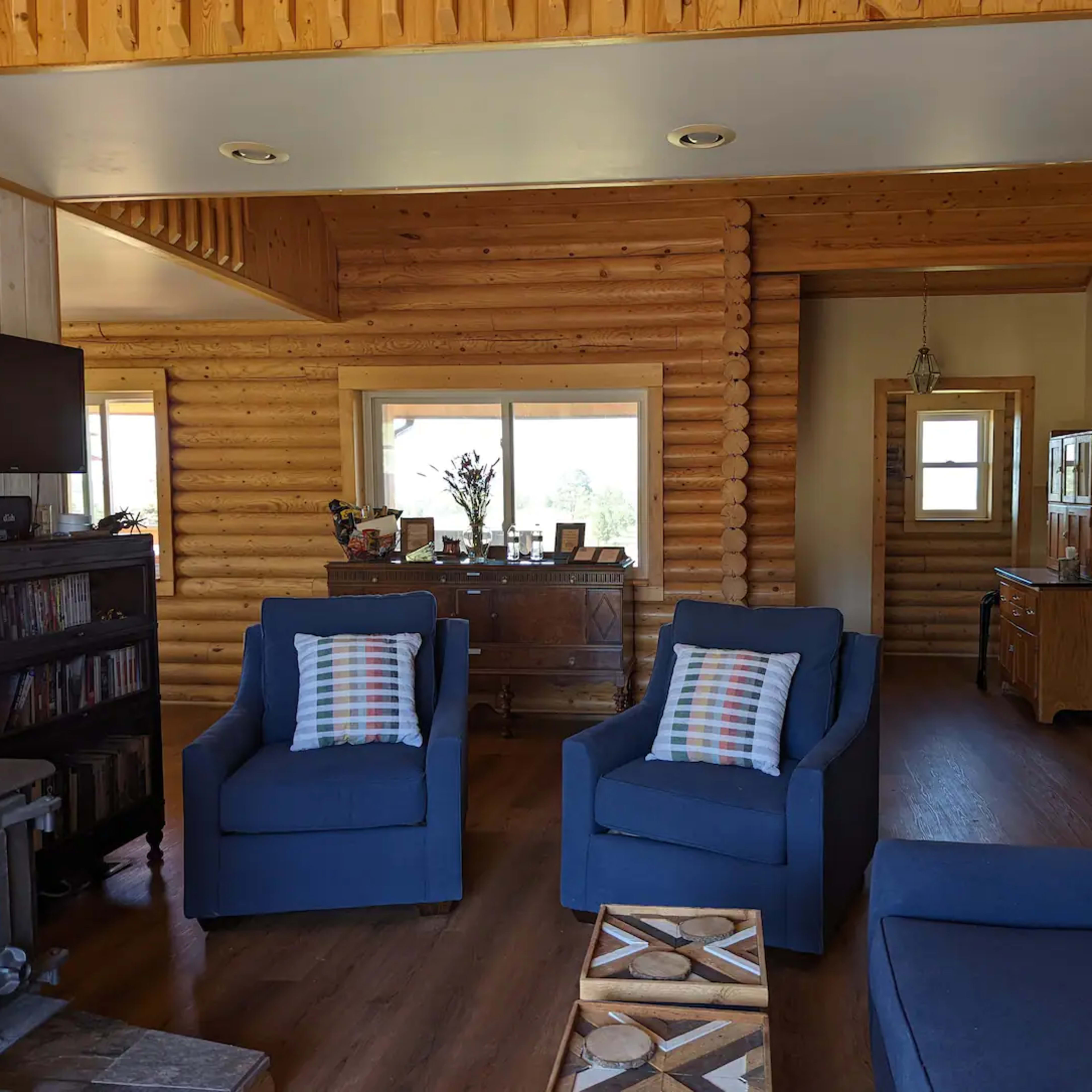 A cozy living room featuring blue armchairs, a wooden ceiling, and a large window with natural light.