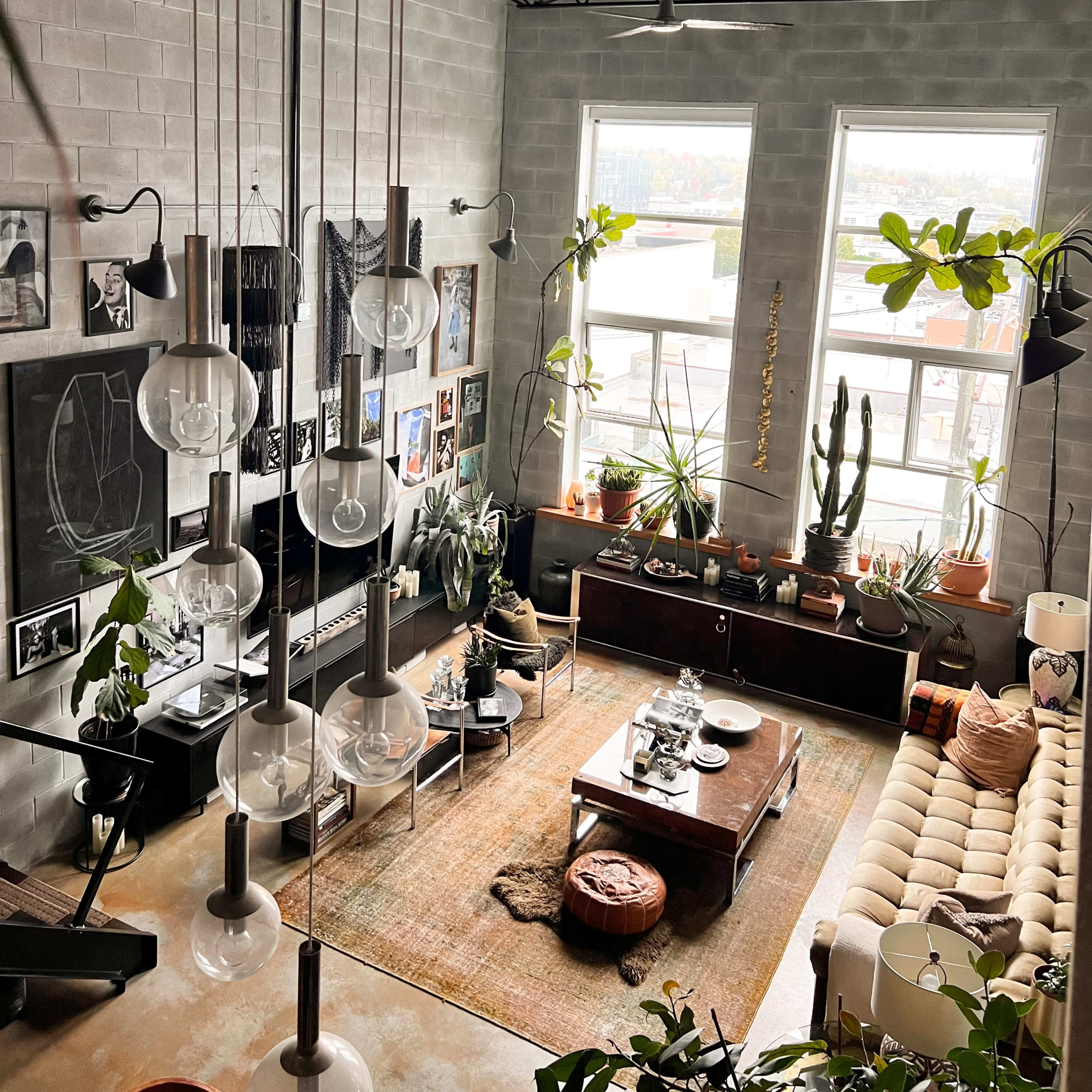 A spacious living room with large windows, various plants, and a mix of furniture including a beige sofa, a wooden coffee table, and decorative artwork on the walls.