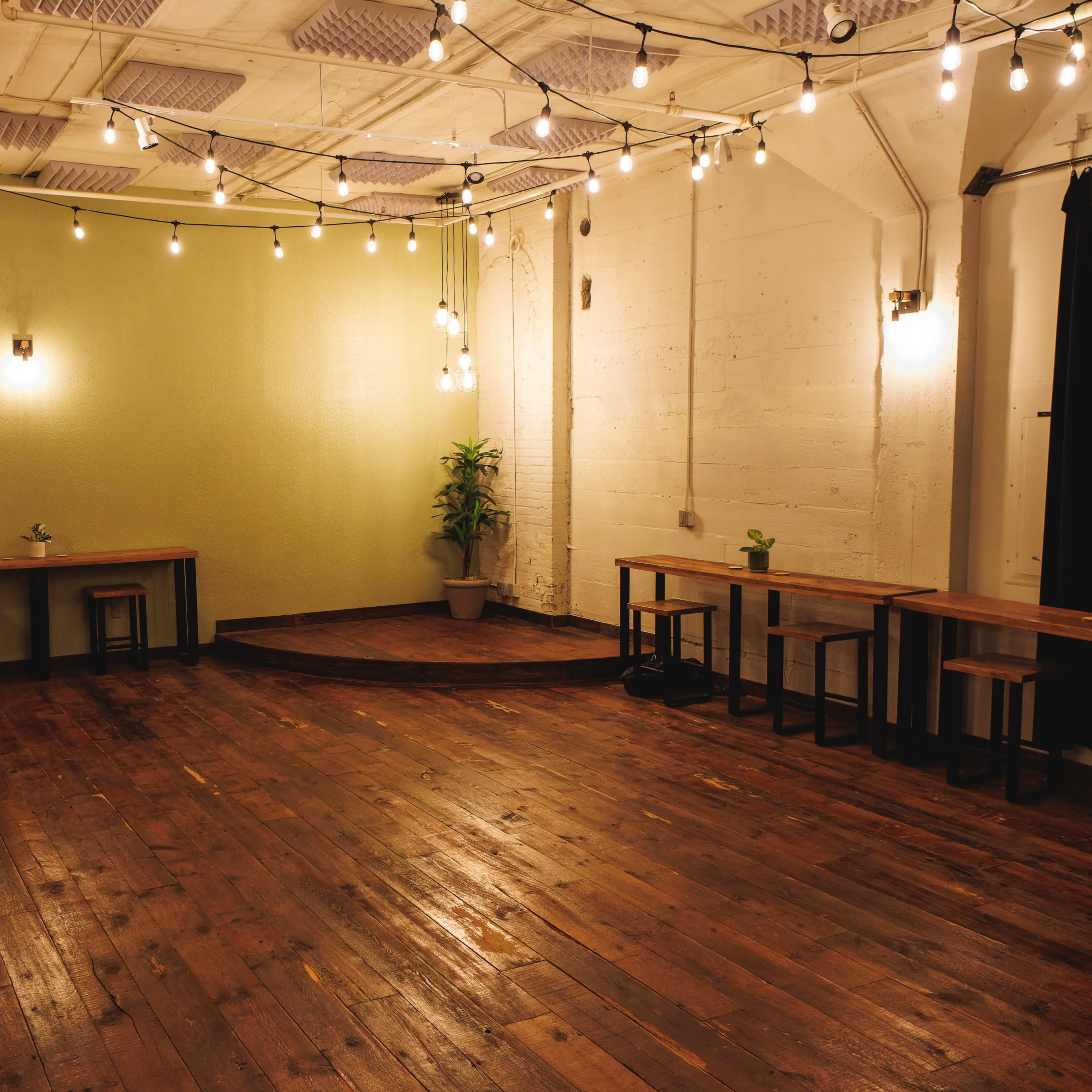 A spacious room with wooden floors, equipped with several wooden tables, plants, and overhead string lights.