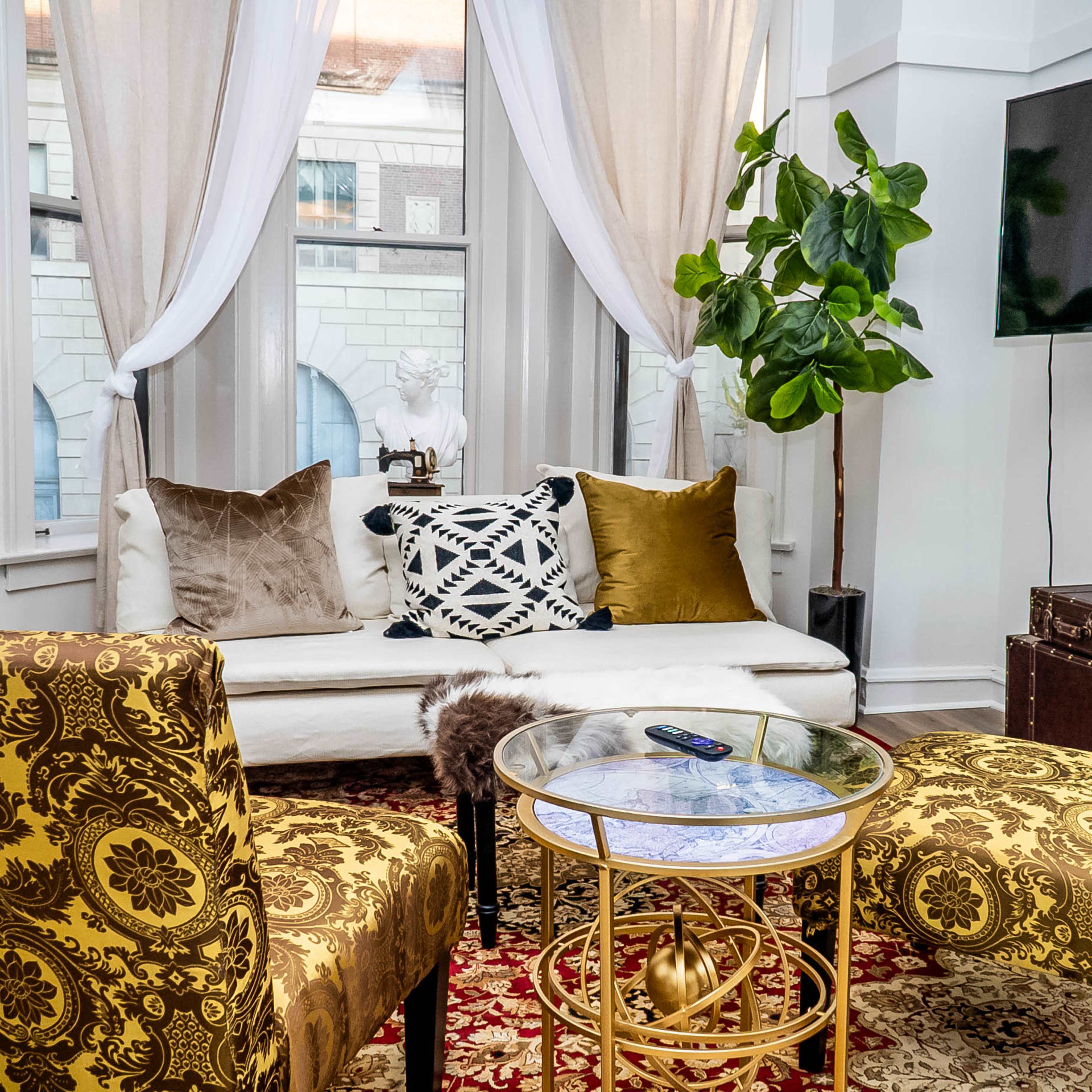 A cozy living room features a white sofa with decorative pillows, two patterned chairs, a glass coffee table, and a television mounted on the wall.