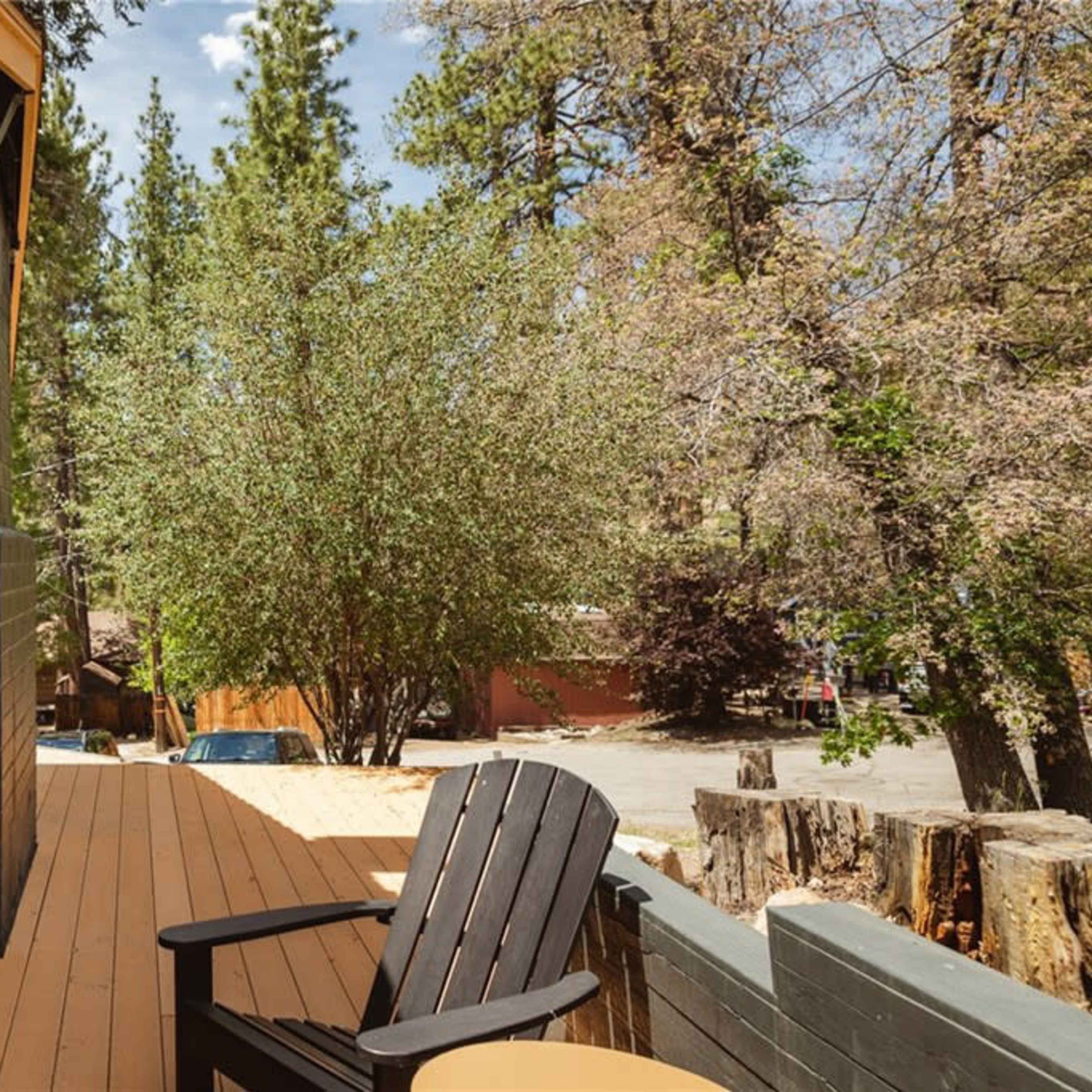 A wooden deck with a black chair overlooking a tree and other greenery amidst tall pine trees.