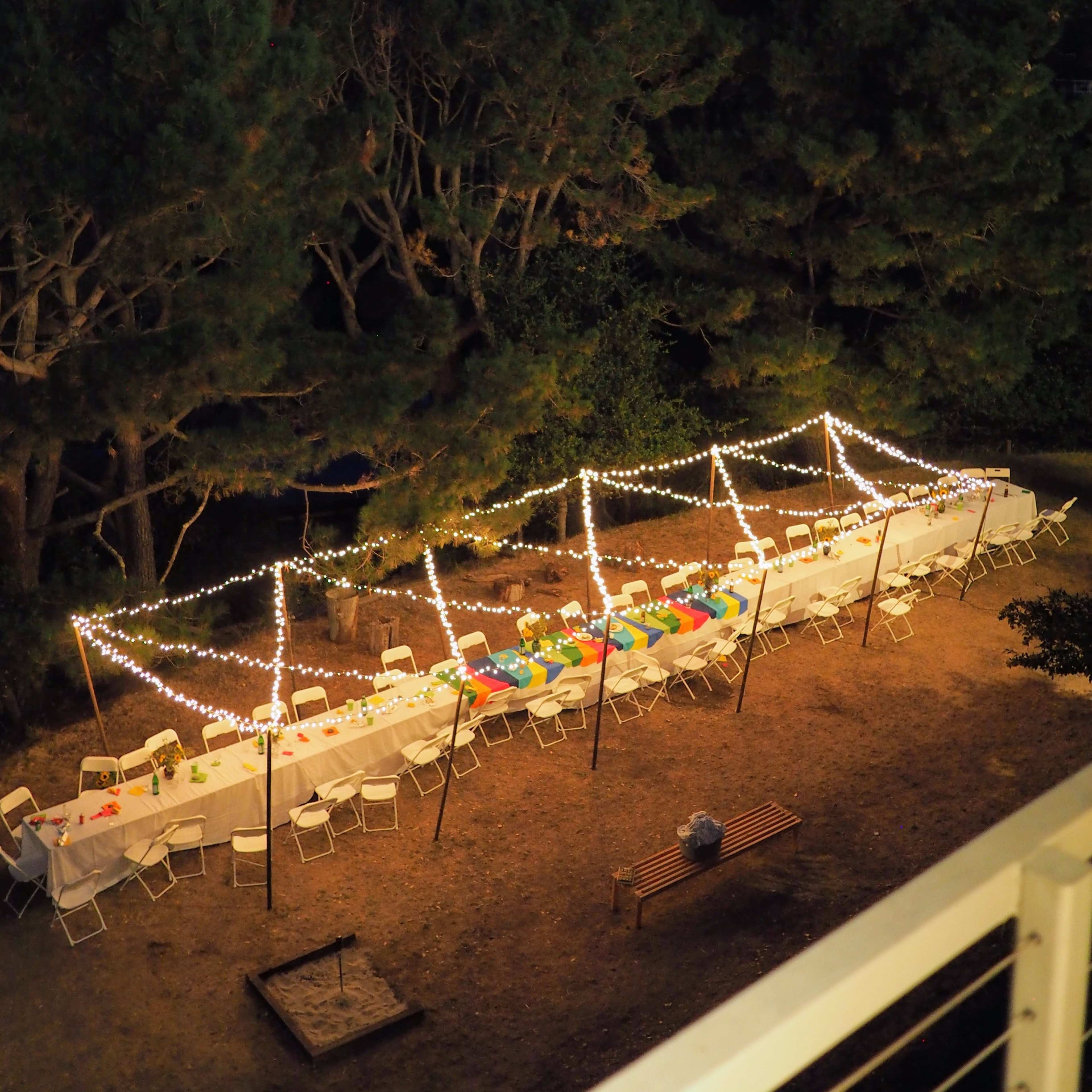 A long dining table with colorful decorations is set up outdoors, surrounded by trees and lit by string lights as dusk falls.