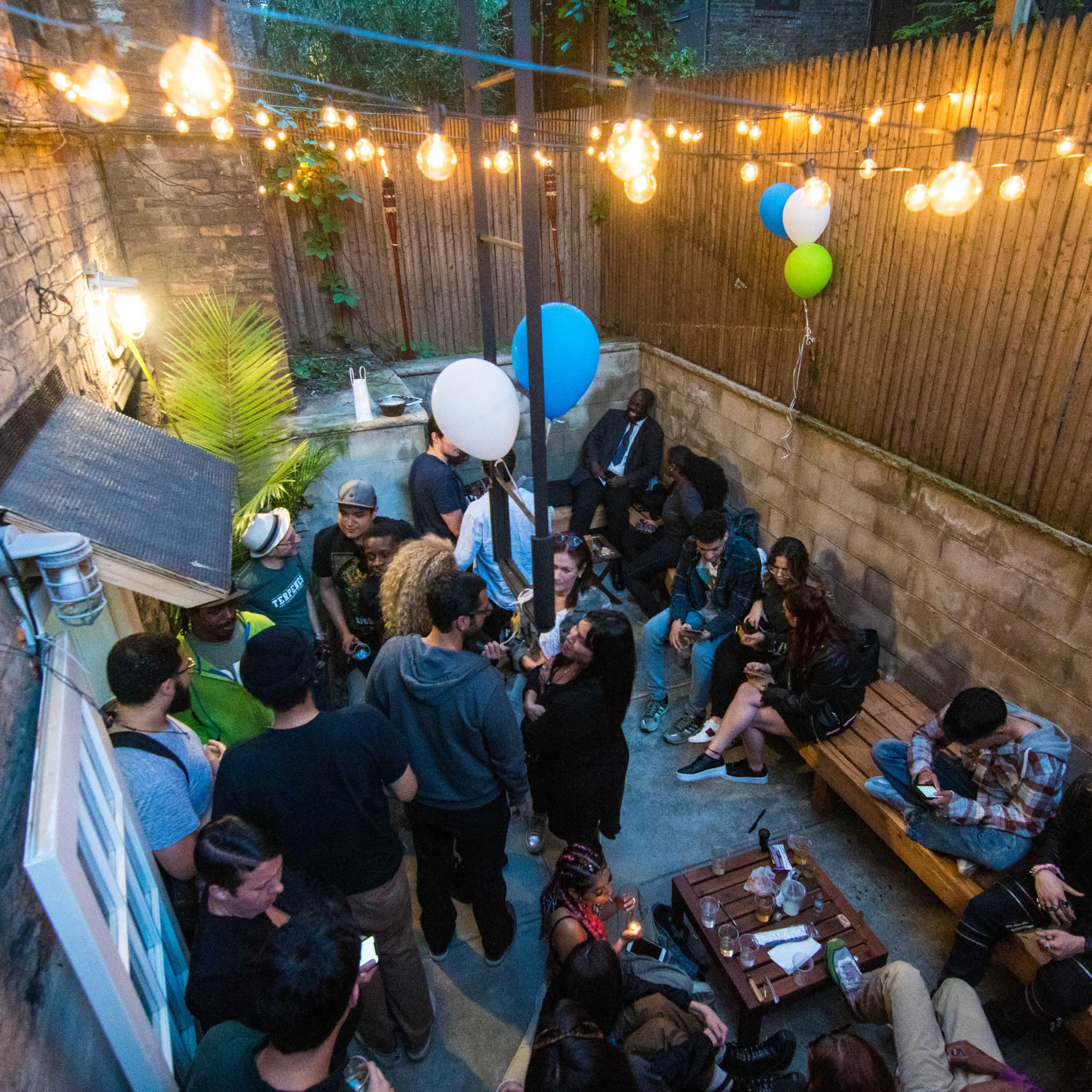 A gathering of people socializes in a dimly lit outdoor patio adorned with string lights and balloons.