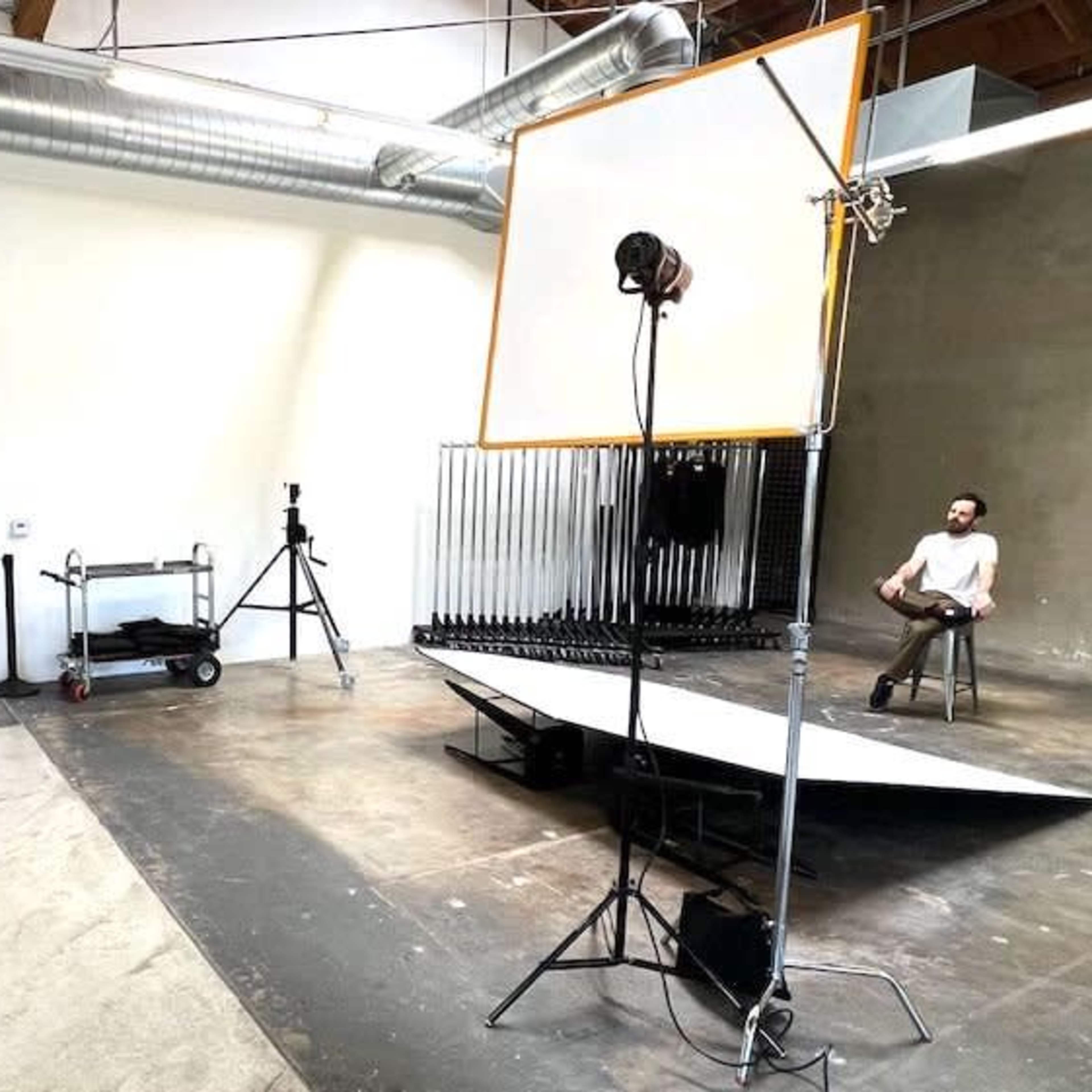 A photographer takes a picture of a seated model in a spacious, industrial-style studio with a large reflector and backdrop.