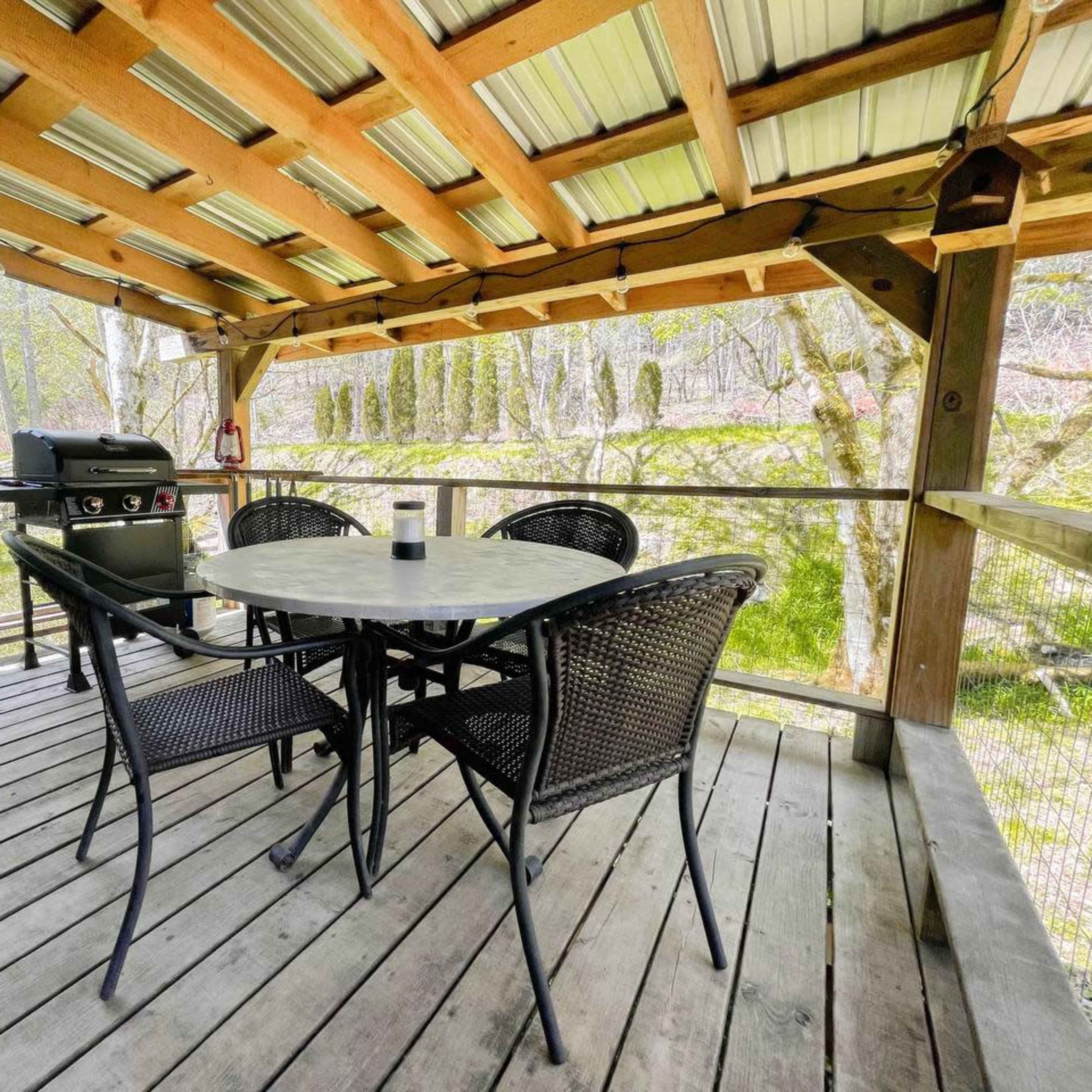 A wooden deck with a round table and four black chairs, alongside a grill and surrounded by trees.