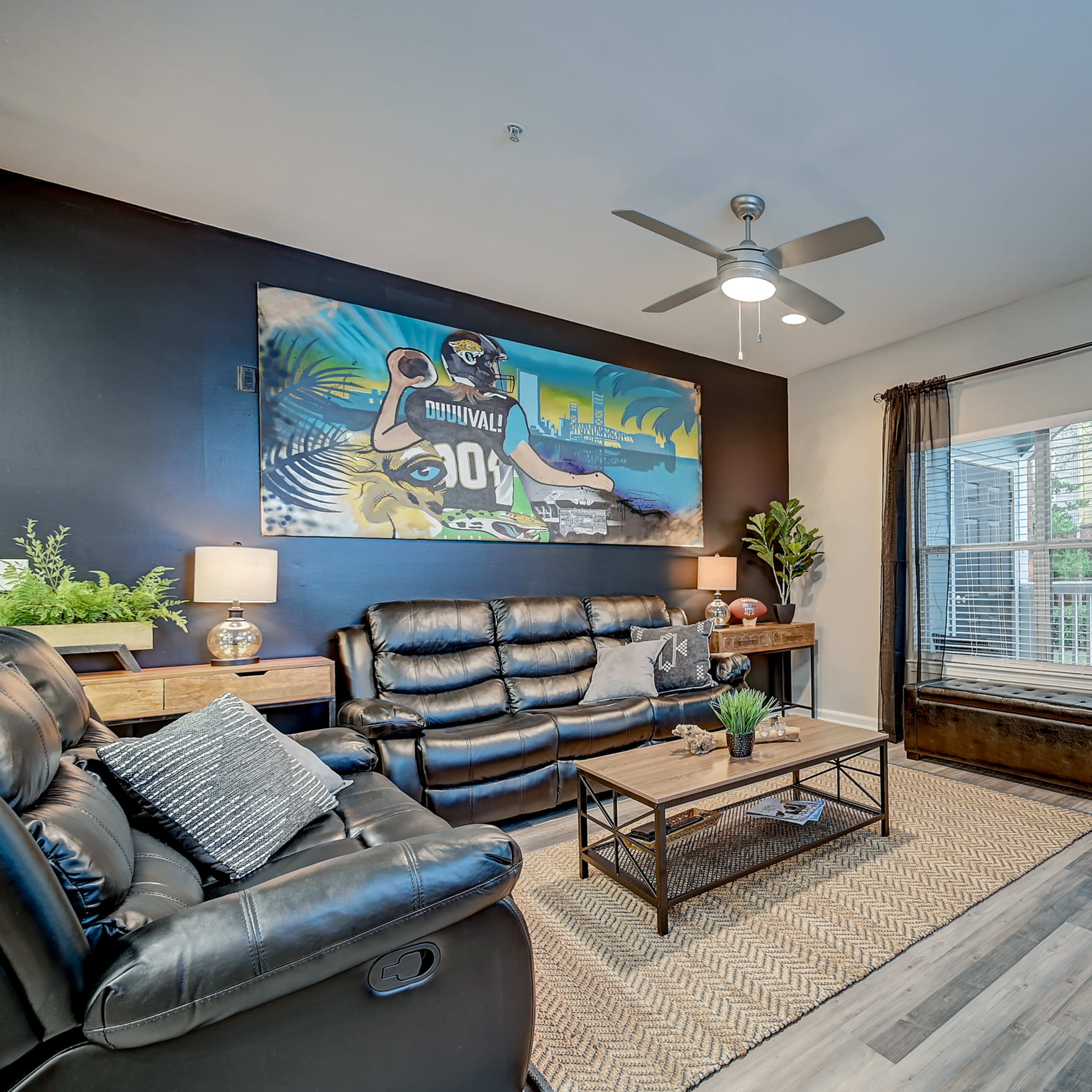 A living room featuring black leather furniture, a wall mural, potted plants, and a patio door with blinds.