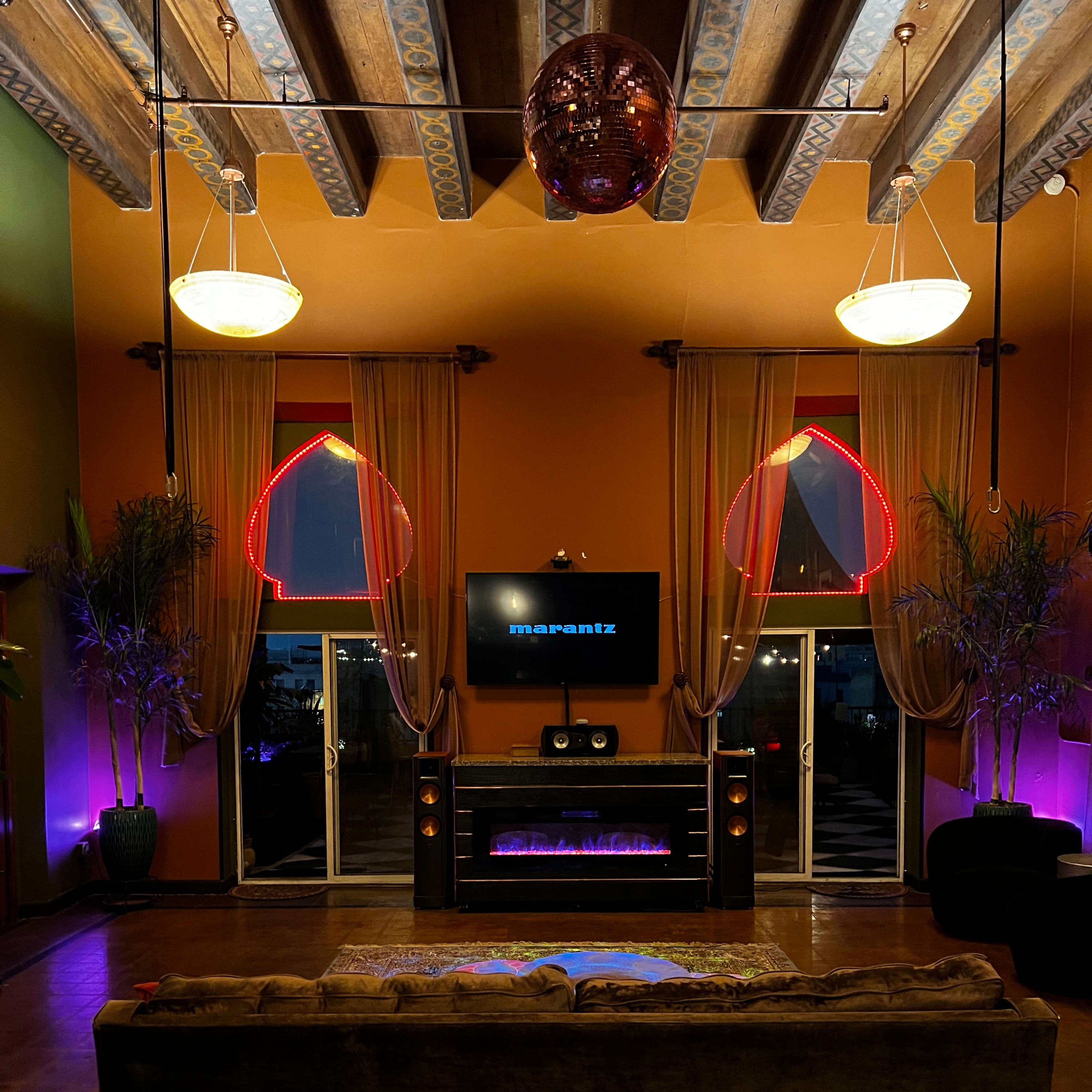 A dimly lit lounge area with a velvet sofa facing a wall-mounted television, framed by large windows illuminated with pink and purple lights.