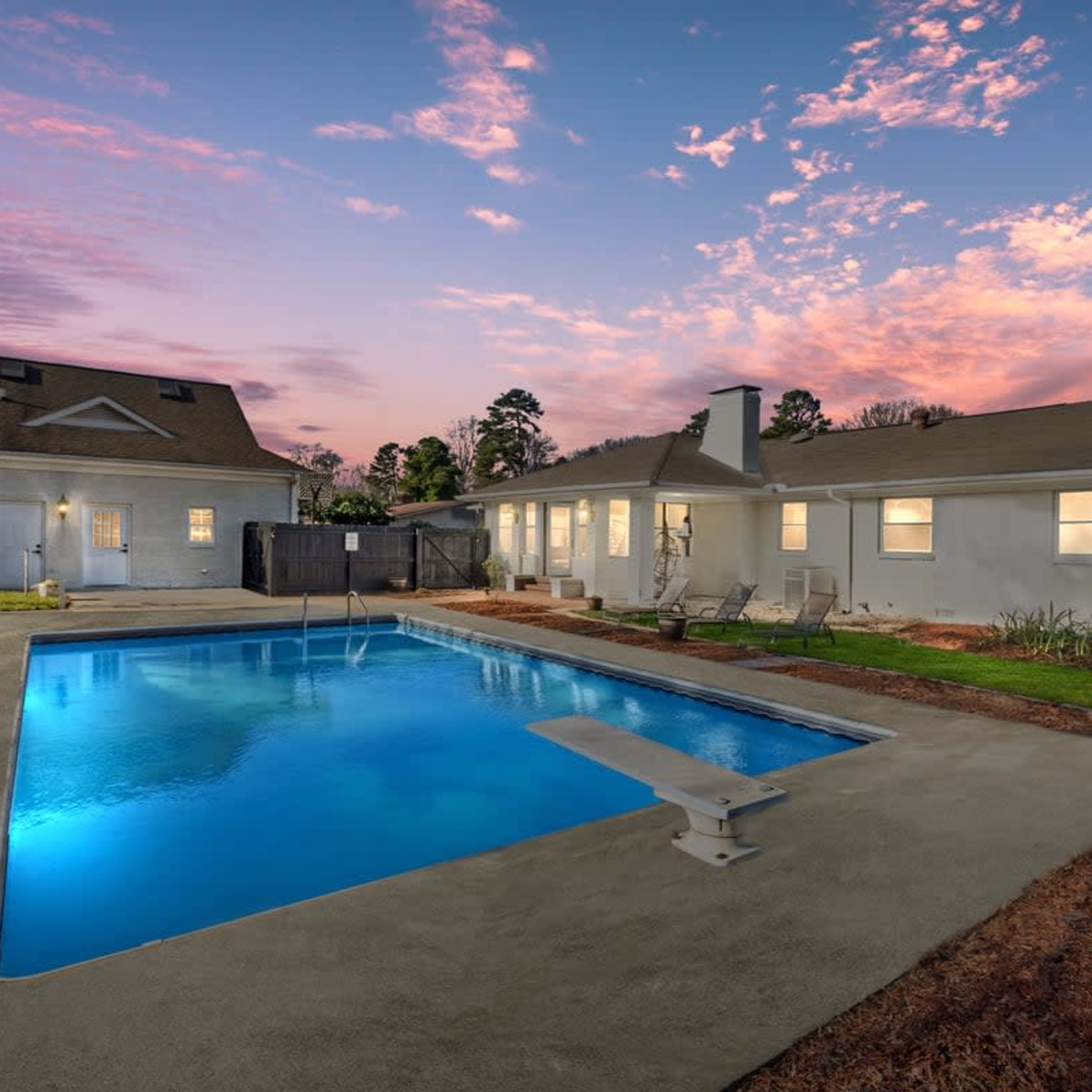 A rectangular swimming pool with a diving board is situated in a backyard, surrounded by grass and flower beds, under a colorful sky at sunset.