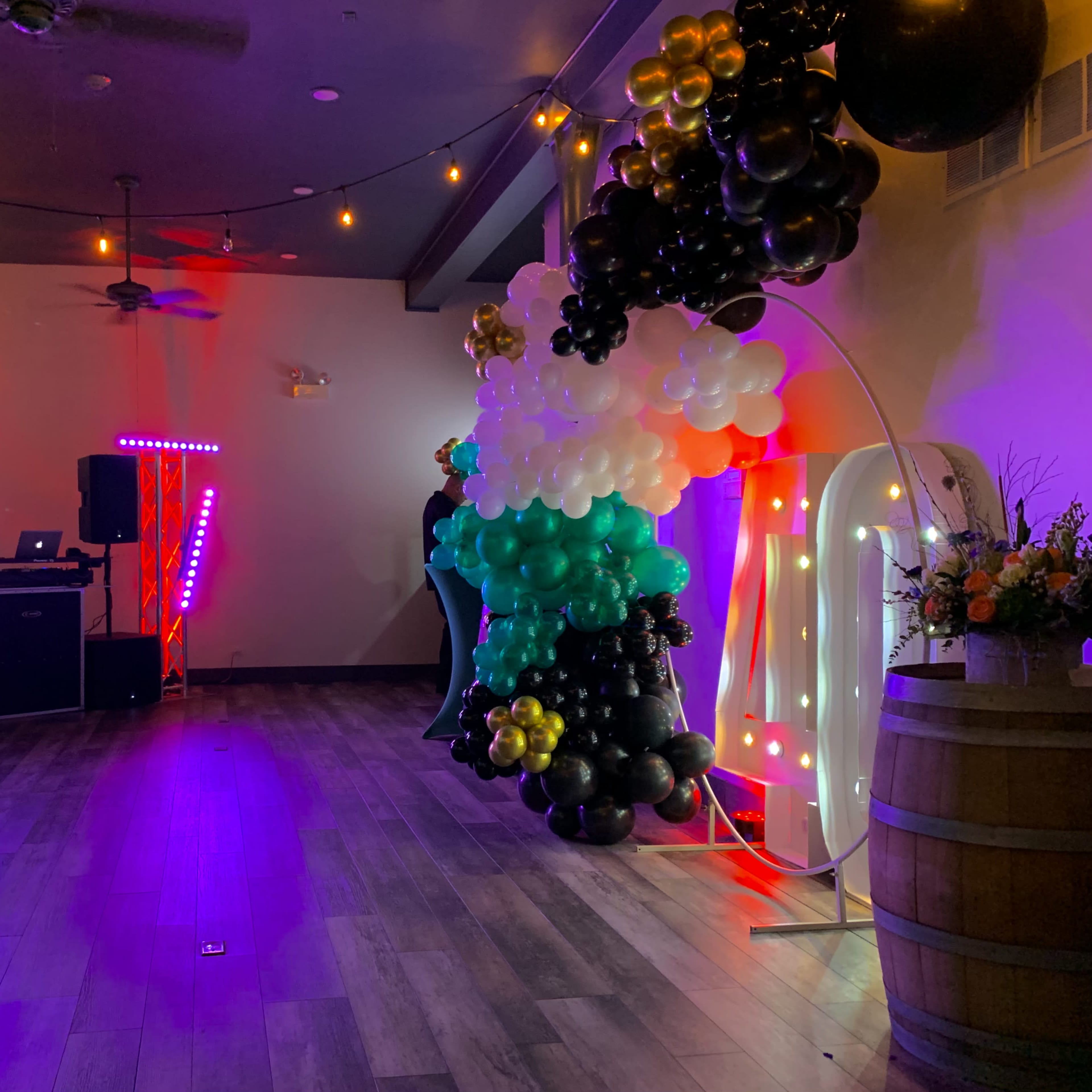 The interior of a venue is decorated with colorful balloons and lights, featuring a DJ setup on one side and a large floral arrangement on a barrel.