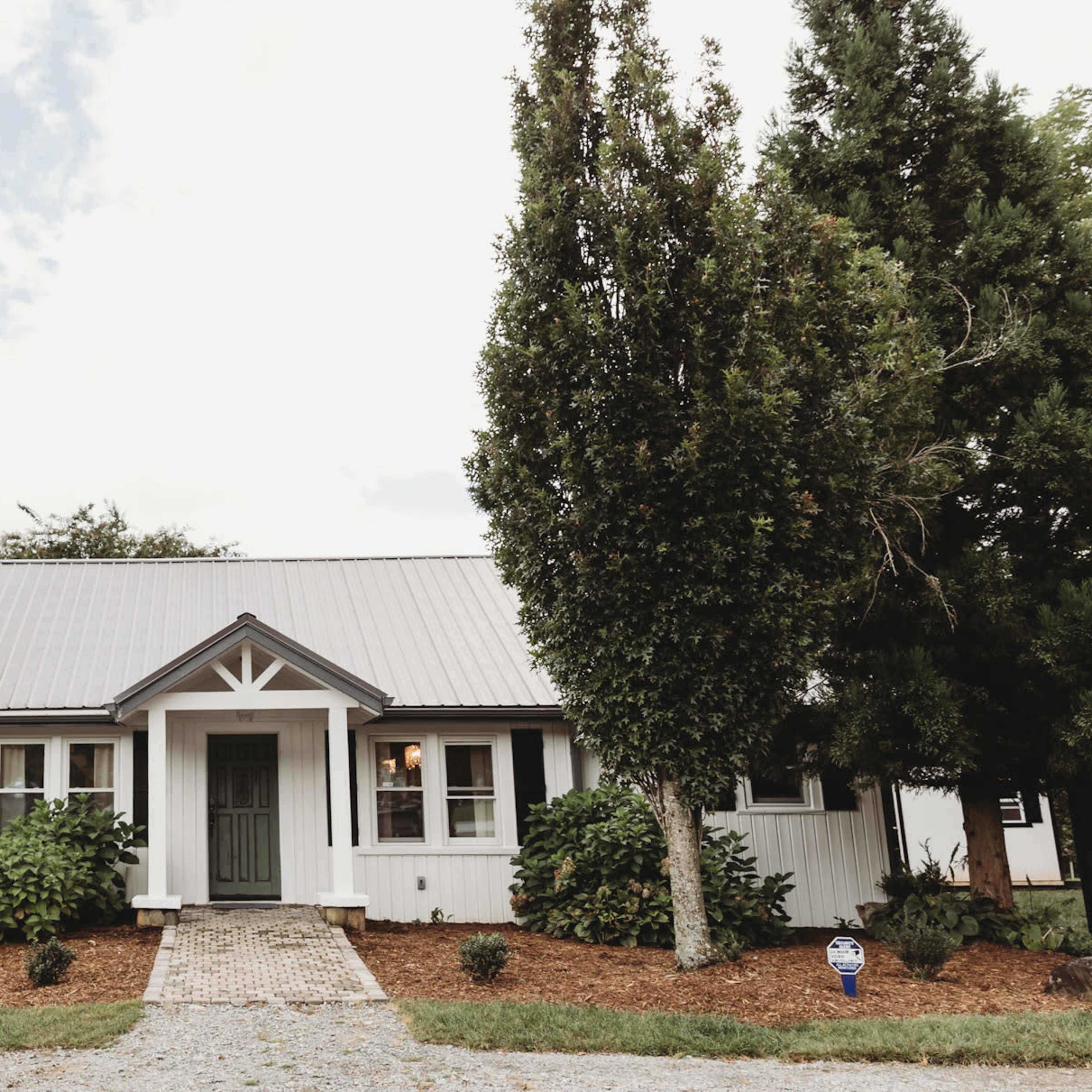 A single-story house with a gray metal roof, surrounded by trees and shrubs, features a central front door framed by two tall conifers.