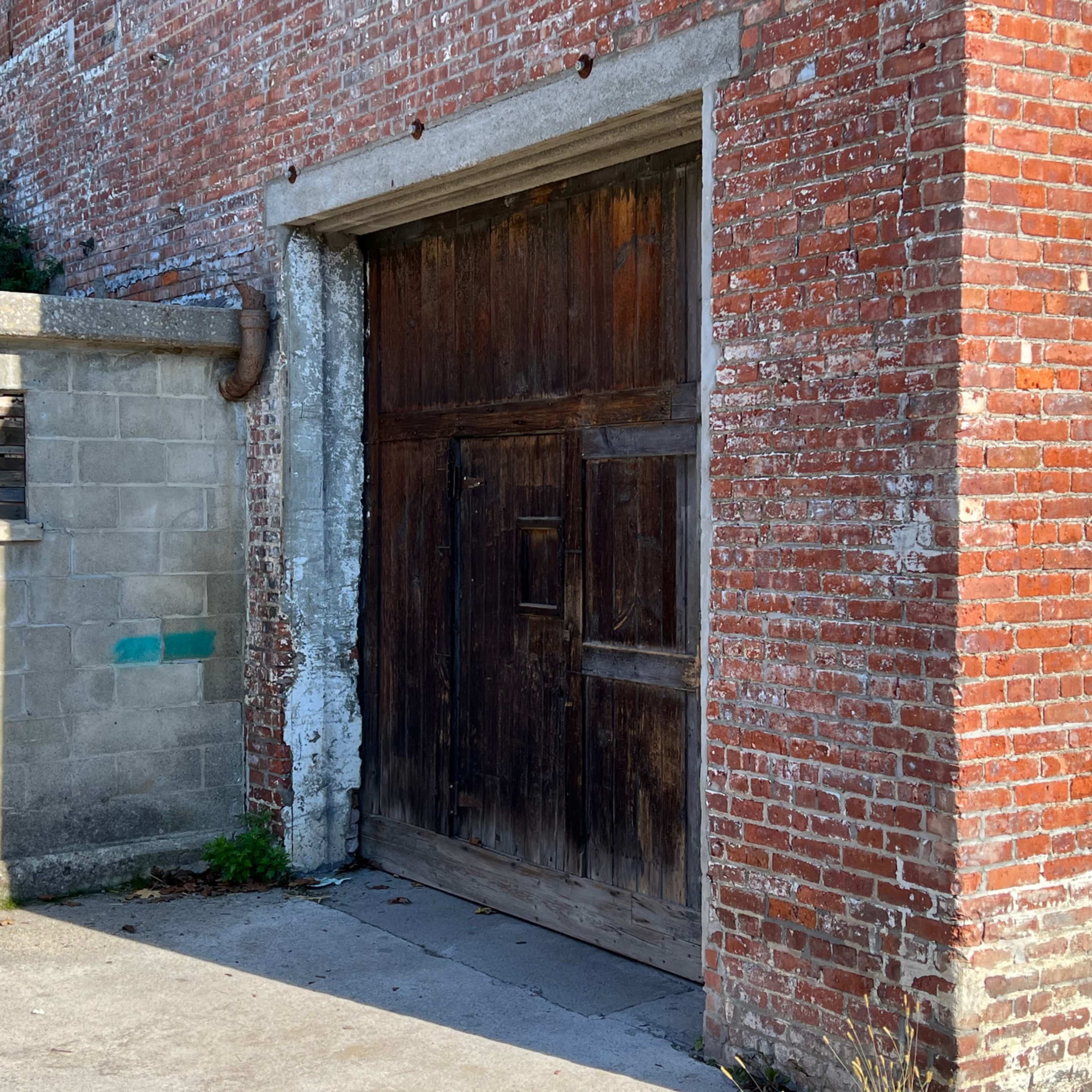 A weathered wooden door embedded within a brick wall, adjacent to a gray concrete structure.