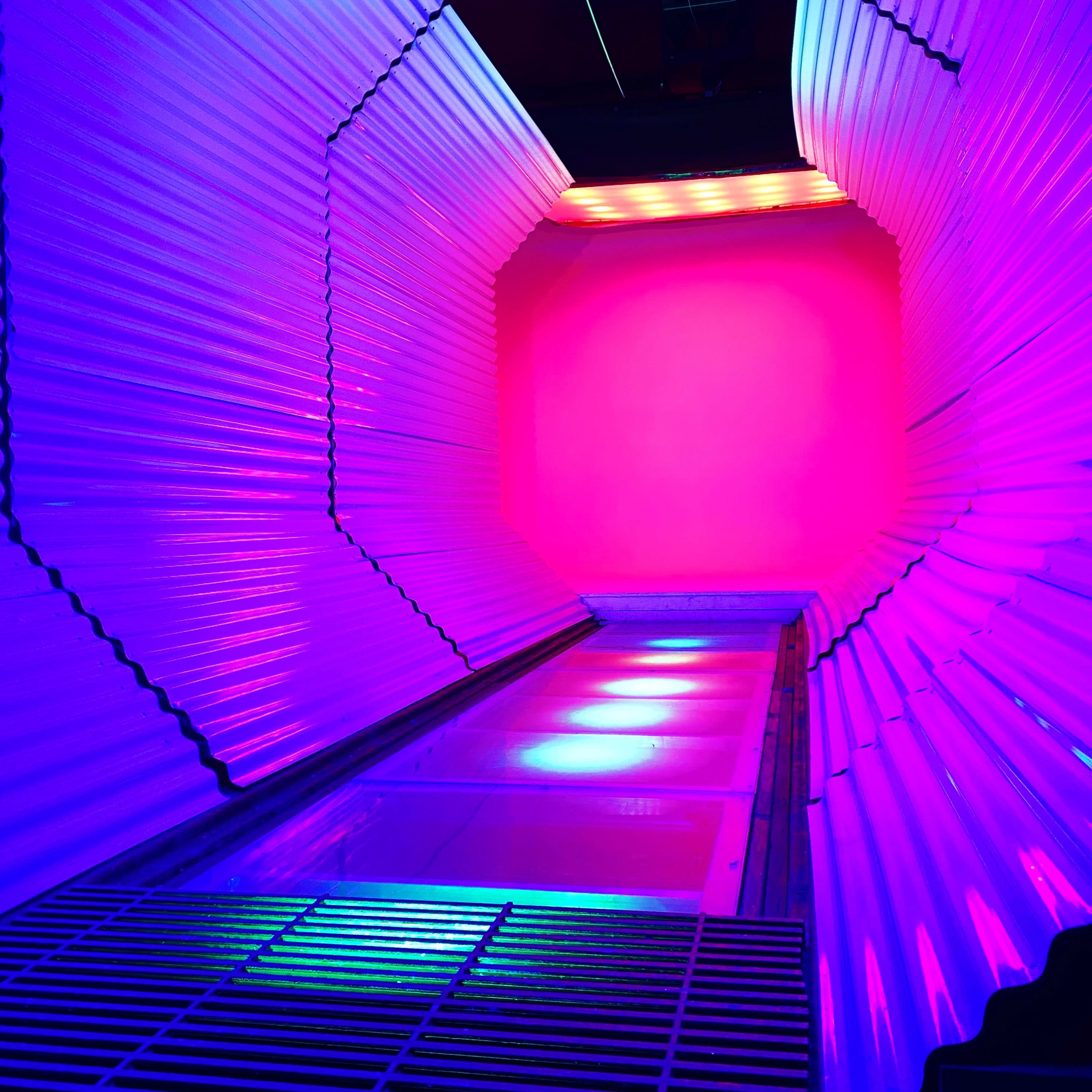 A brightly lit corridor with curved metallic walls and a vibrant pink and blue color scheme, leading to a solid wall at the end.