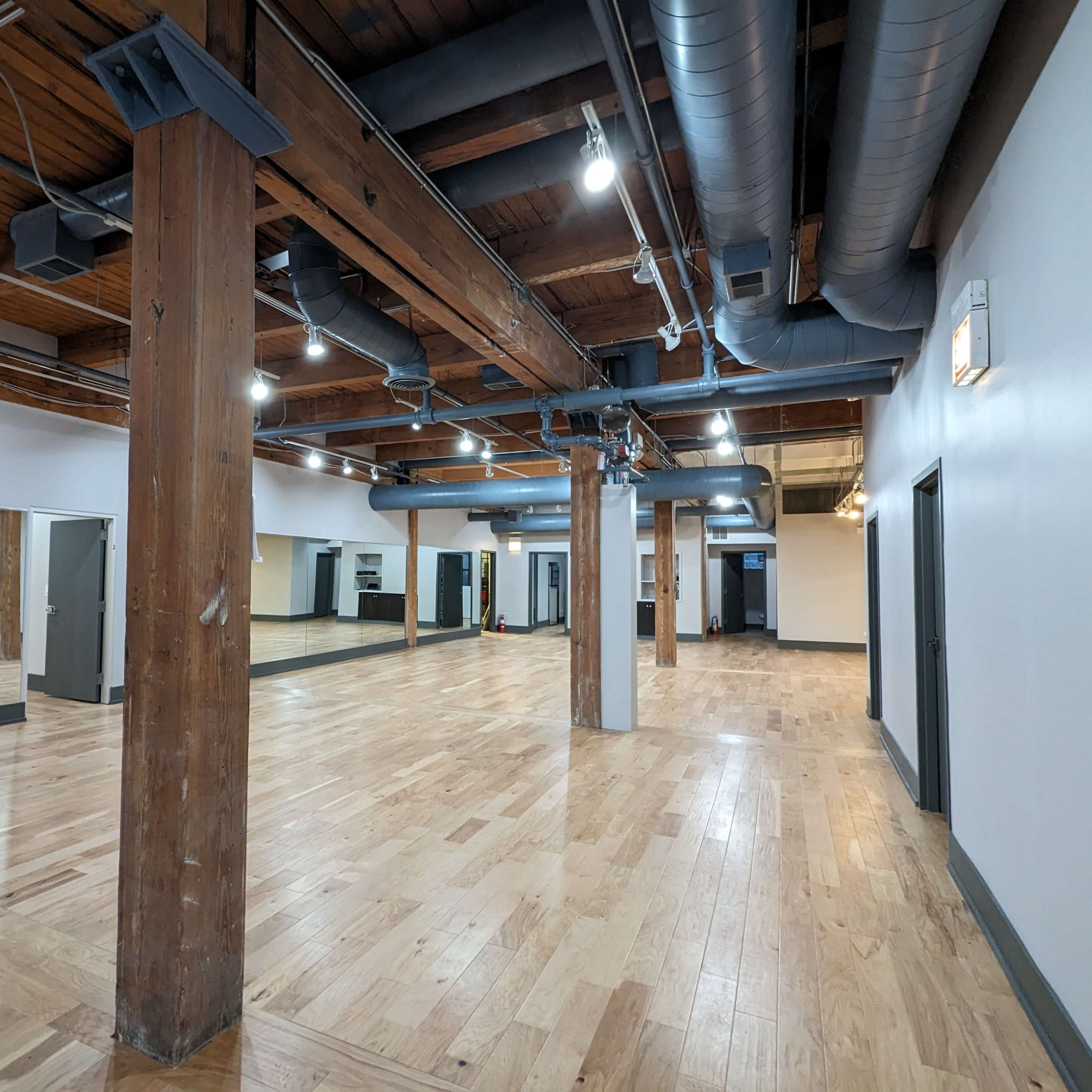 A spacious, empty wooden-floored studio with exposed overhead beams and large mirrors on one wall.