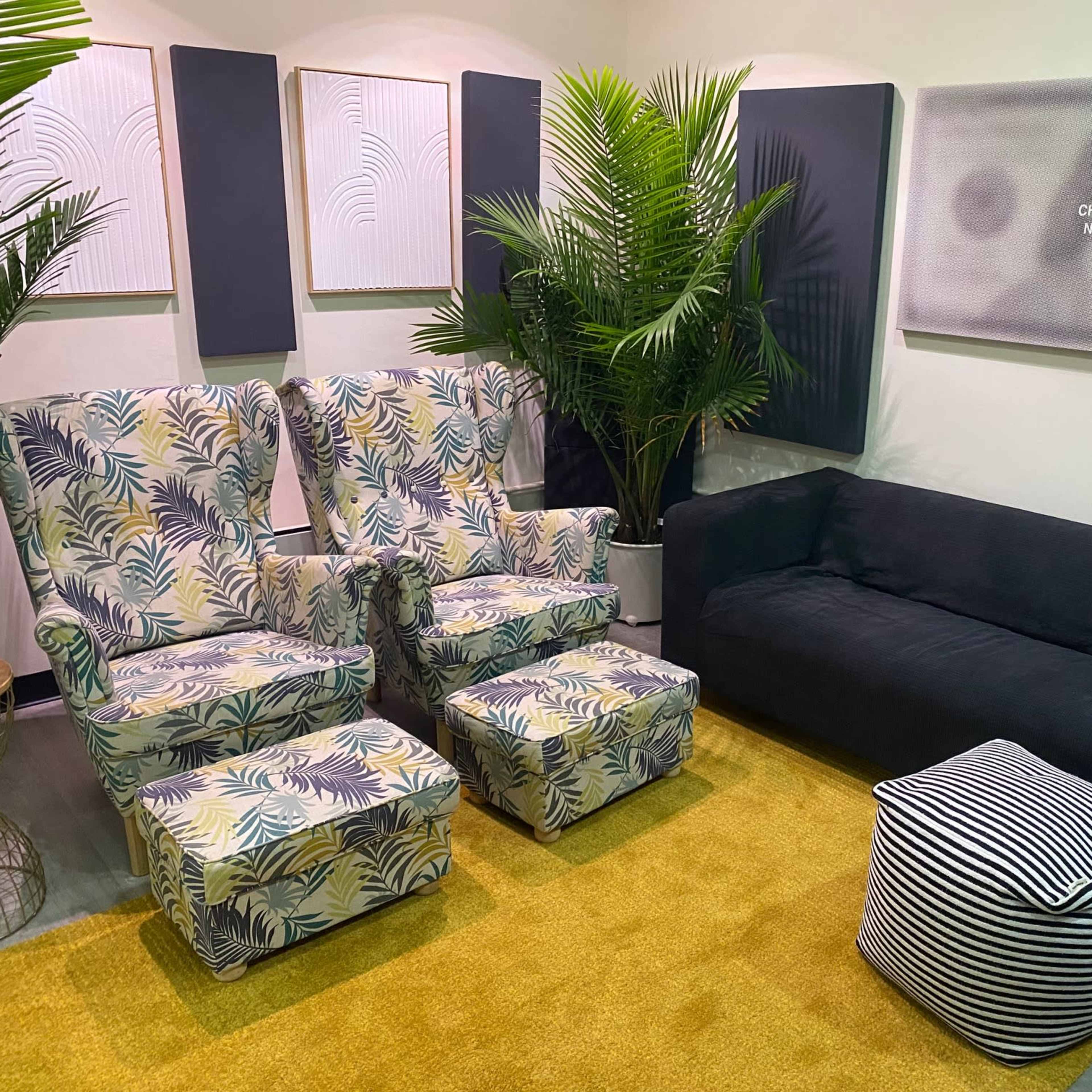 A cozy seating area featuring two patterned armchairs, a dark couch, a striped pouf, and tropical plants on a yellow rug.