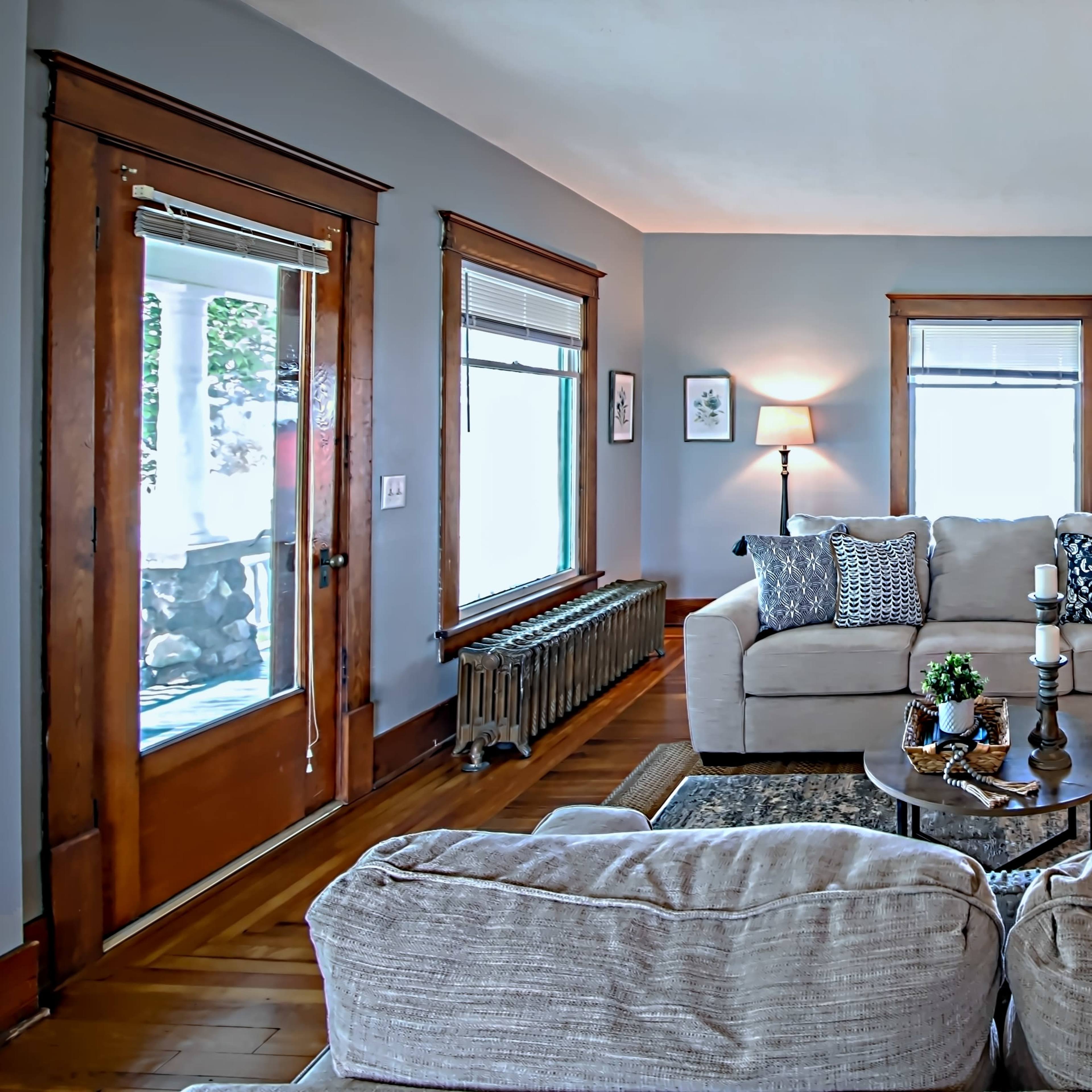 A cozy living room with a light-colored couch, wooden accents, and large windows allowing natural light to enter the space.
