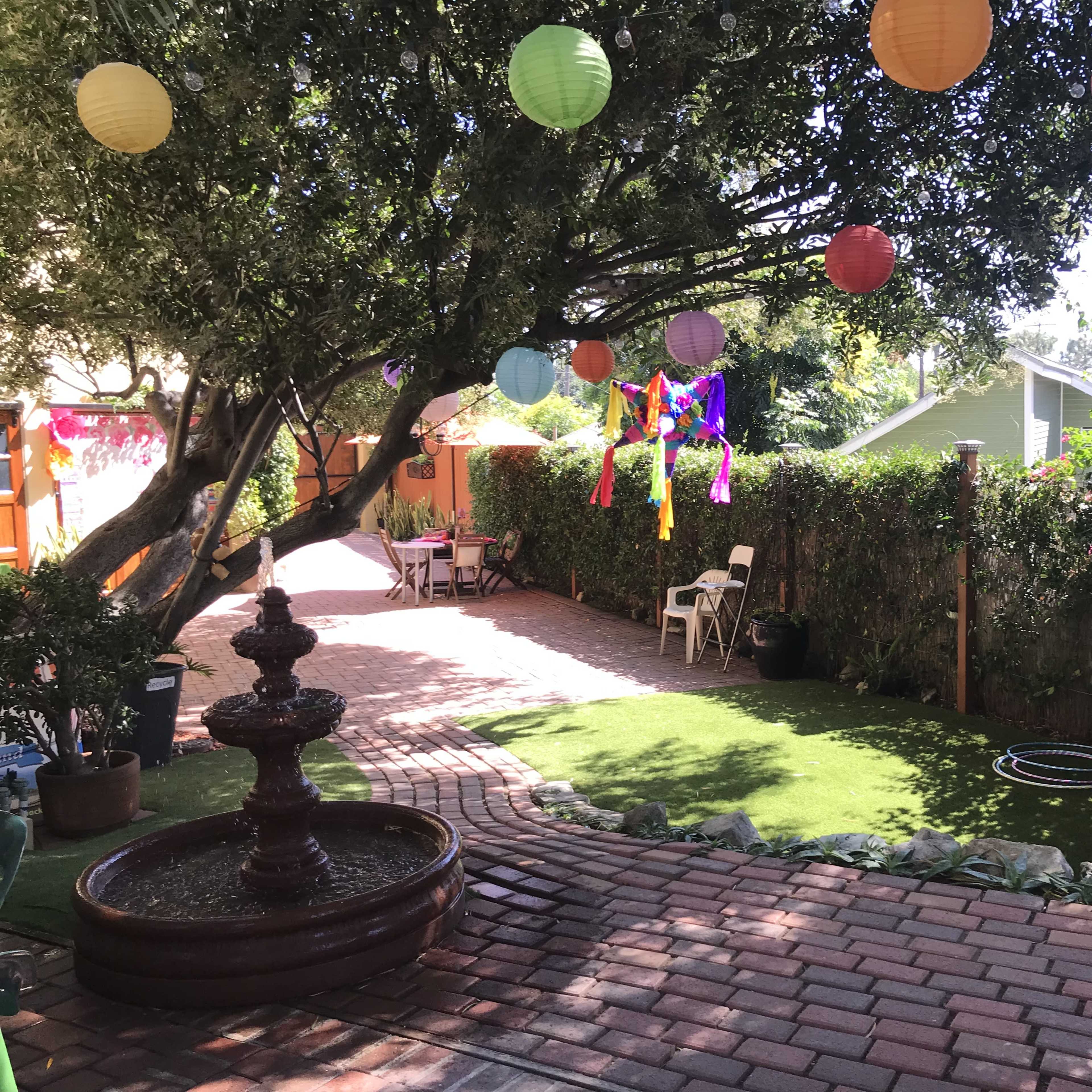 A backyard with a fountain, colorful hanging lanterns, decorated trees, and a pathway lined with bricks and greenery.