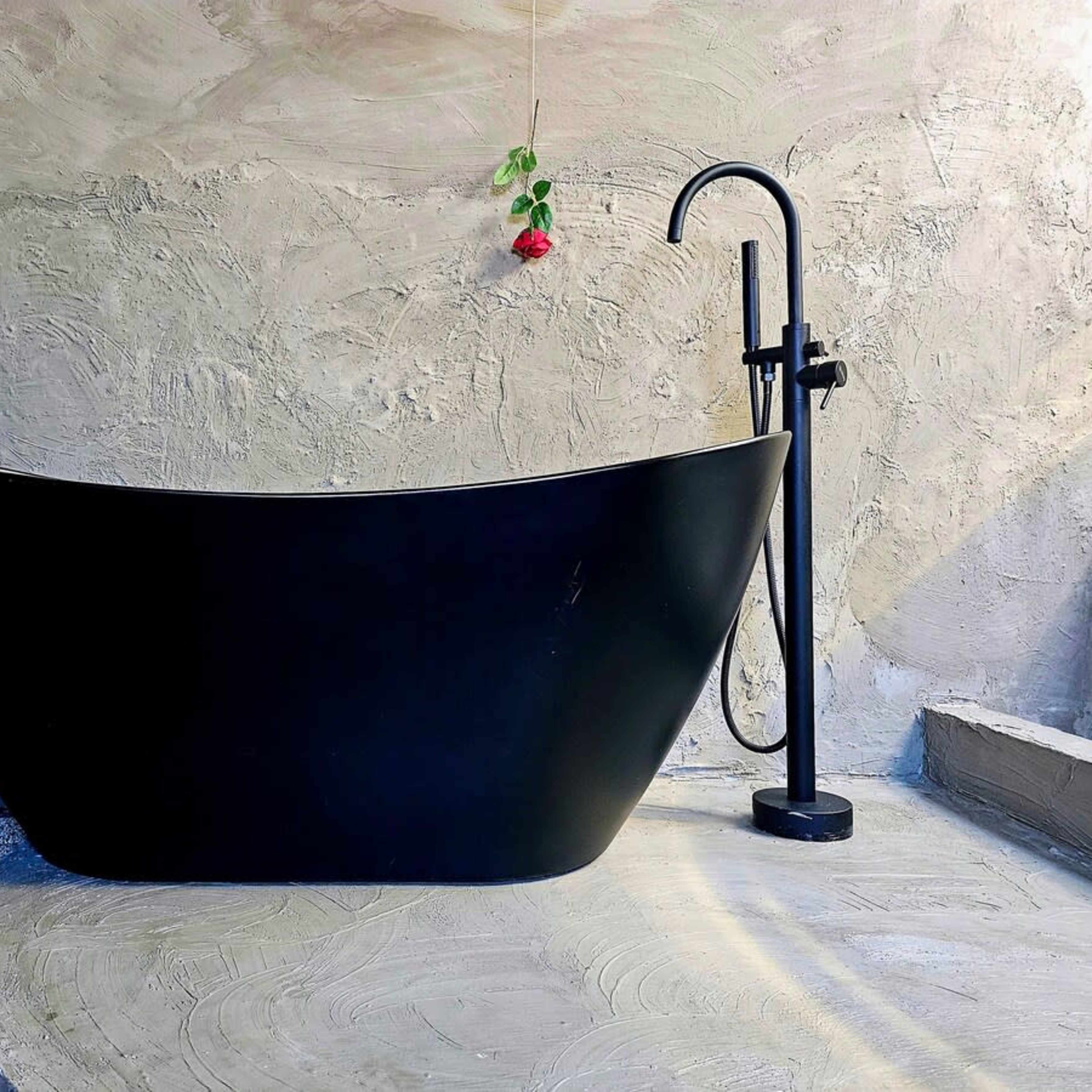 A modern black bathtub on a textured concrete surface, accompanied by a floor-mounted faucet and greenery in the background.