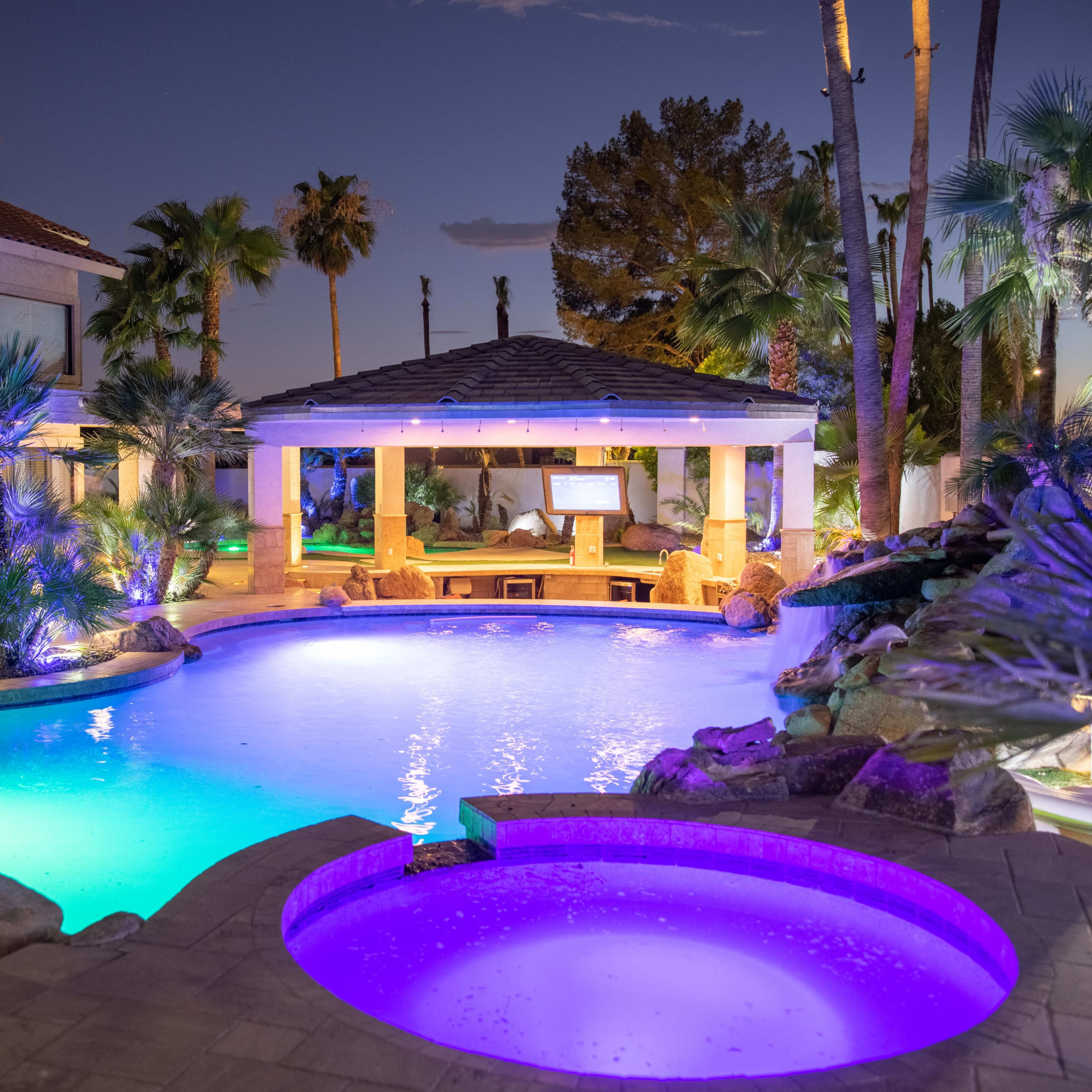 A luxurious backyard pool area with palm trees, a spa, and a covered lounge space, illuminated by colorful lights during twilight.