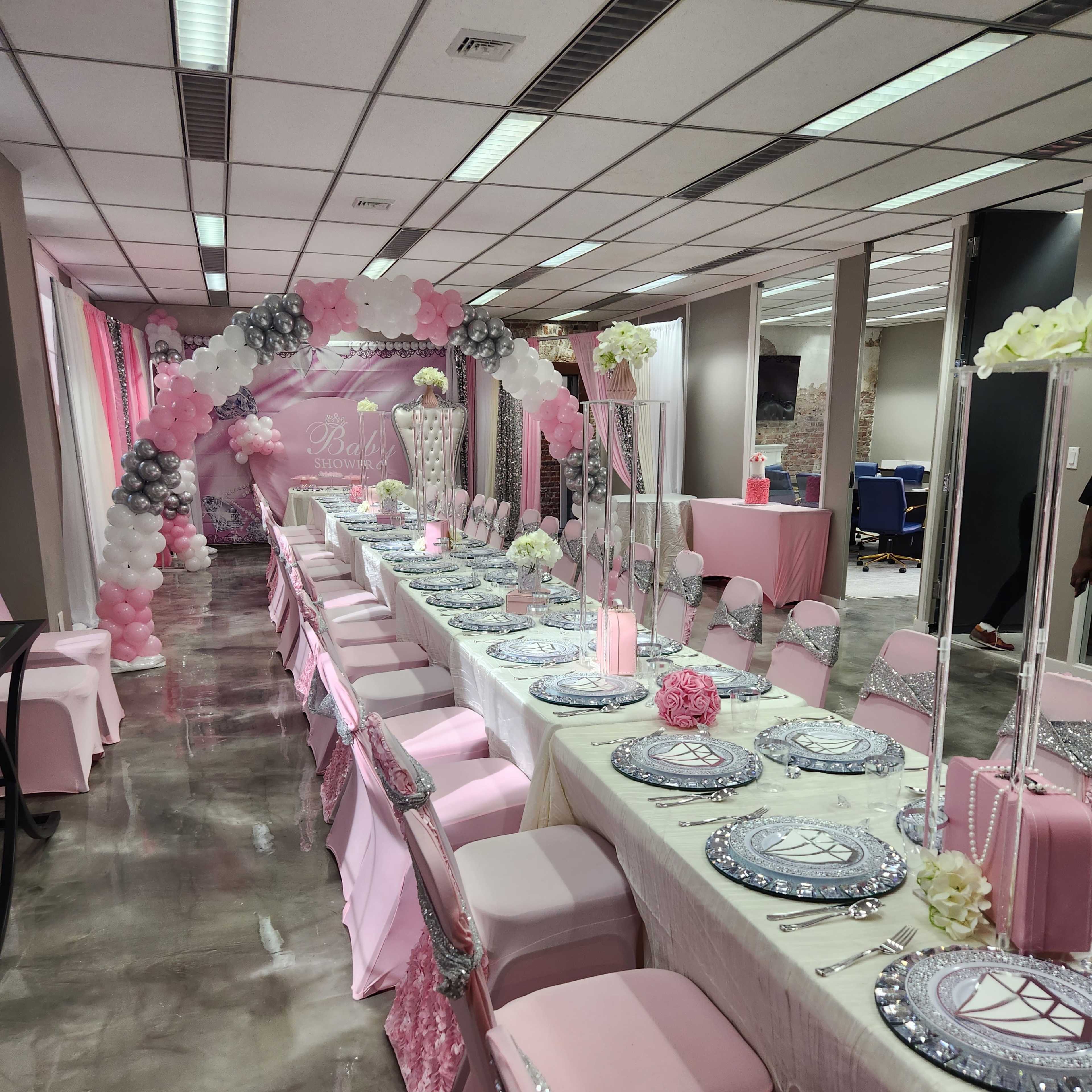 A decorated event space with a long table set for a celebration, featuring pink and silver decor, floral centerpieces, and a balloon arch in the background.