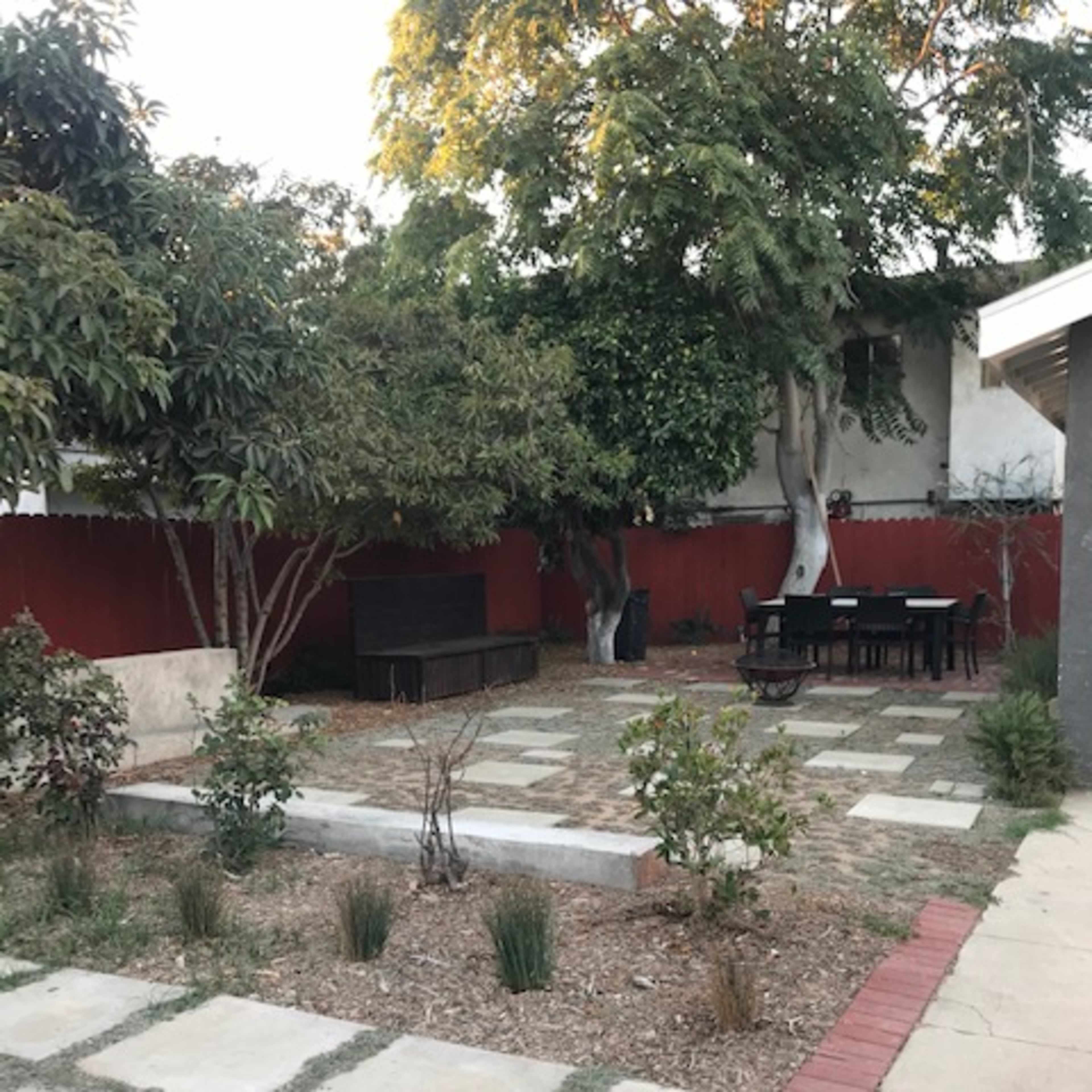 A landscaped backyard with stone pathways, minimal vegetation, a patio set, and a wooden bench near a red fence.
