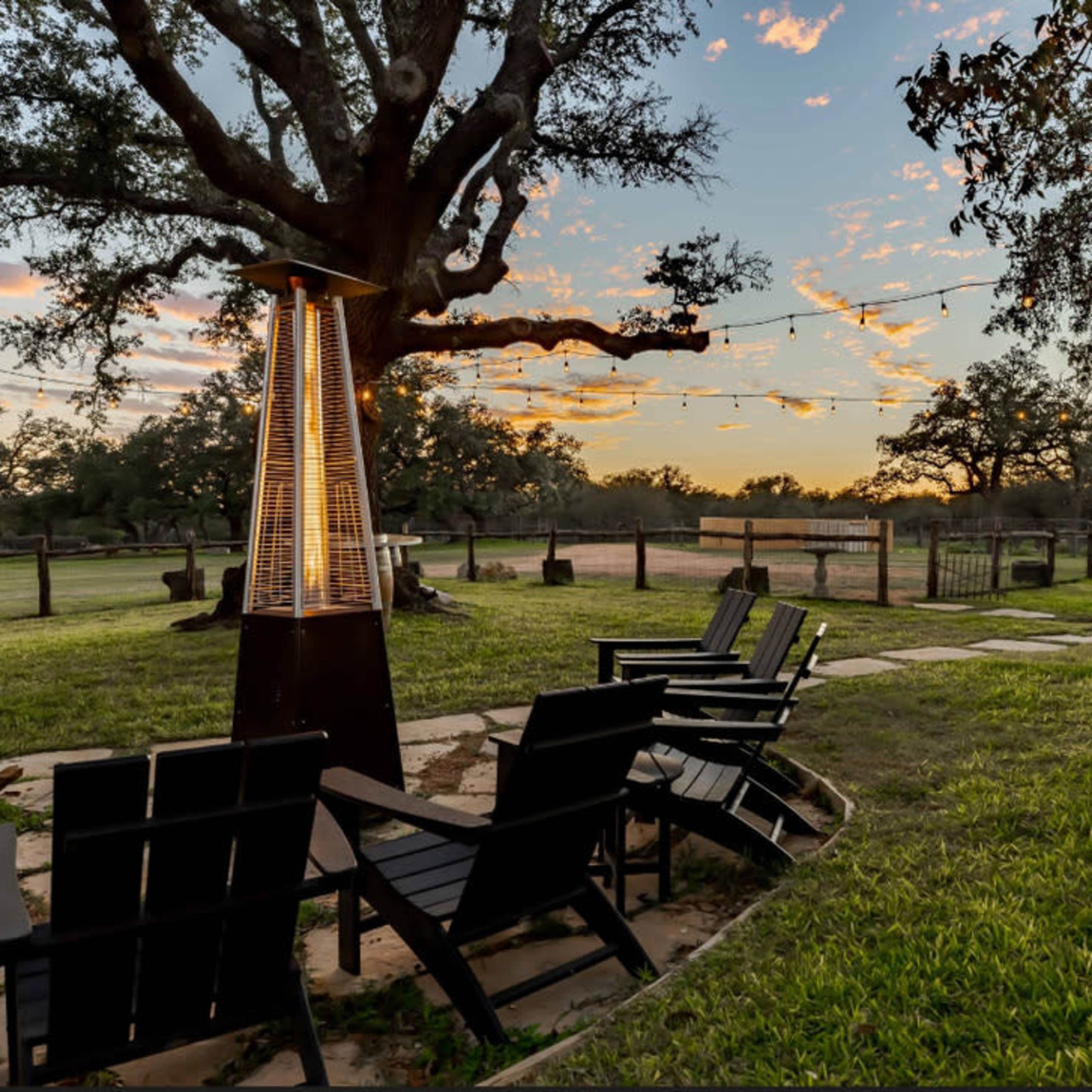 The scene shows a landscaped outdoor area with four chairs arranged around a heater next to a large tree at sunset, with string lights overhead and a grassy field in the background.