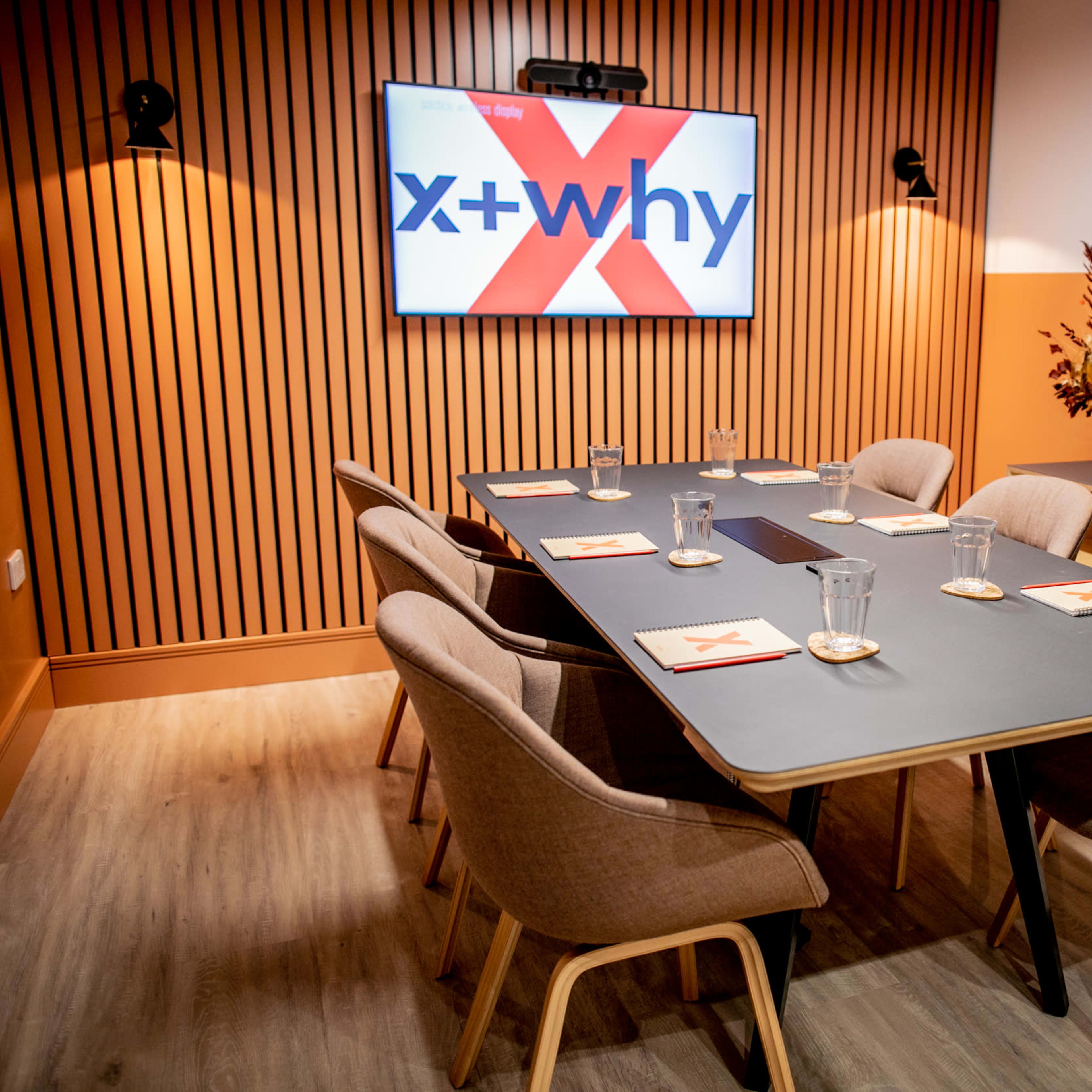 A modern conference room featuring a large table surrounded by six chairs, with a screen displaying "x+why" on the wall and decorative elements in the background.