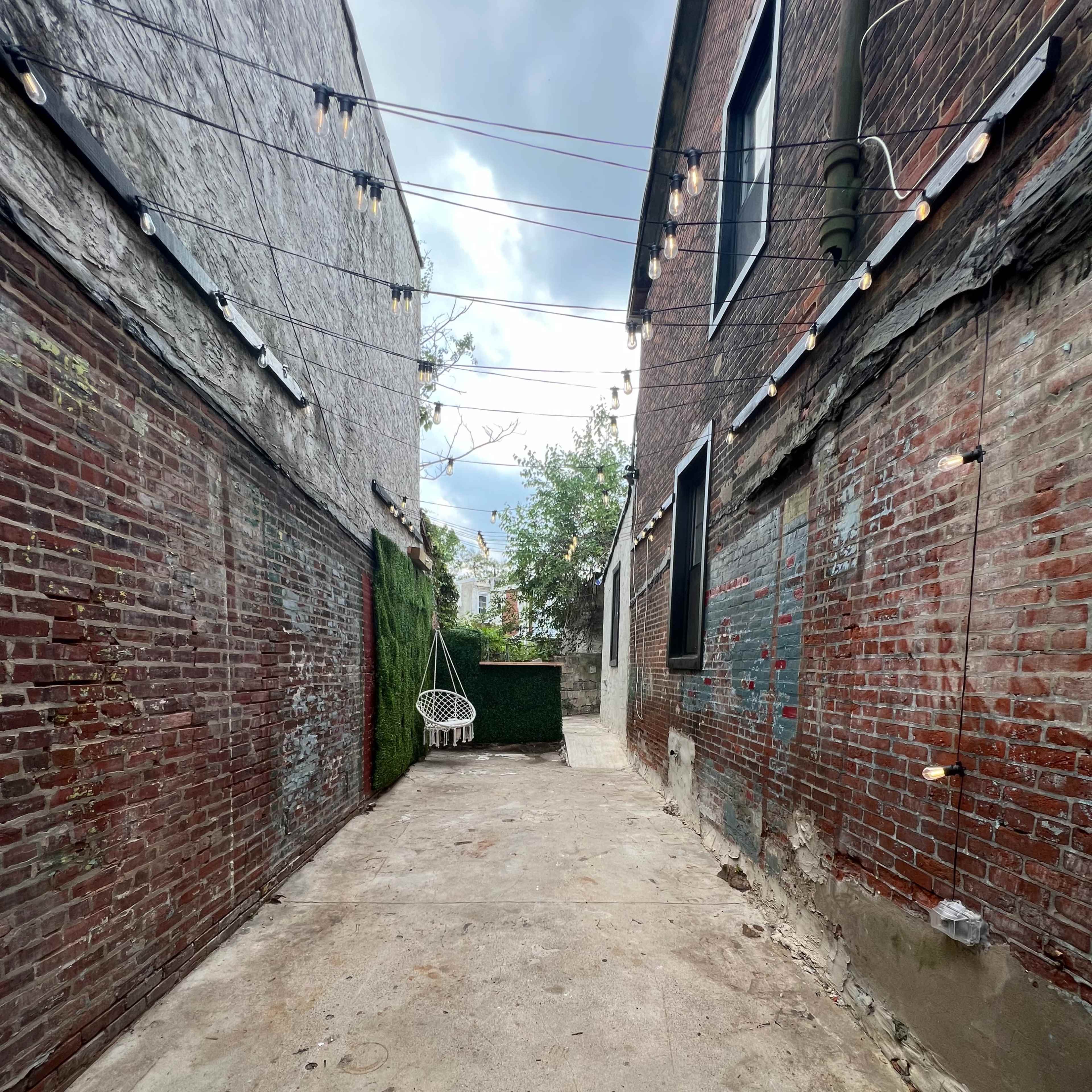 A narrow alleyway flanked by brick walls, leading to a concrete space with a hanging chair and greenery at the far end.