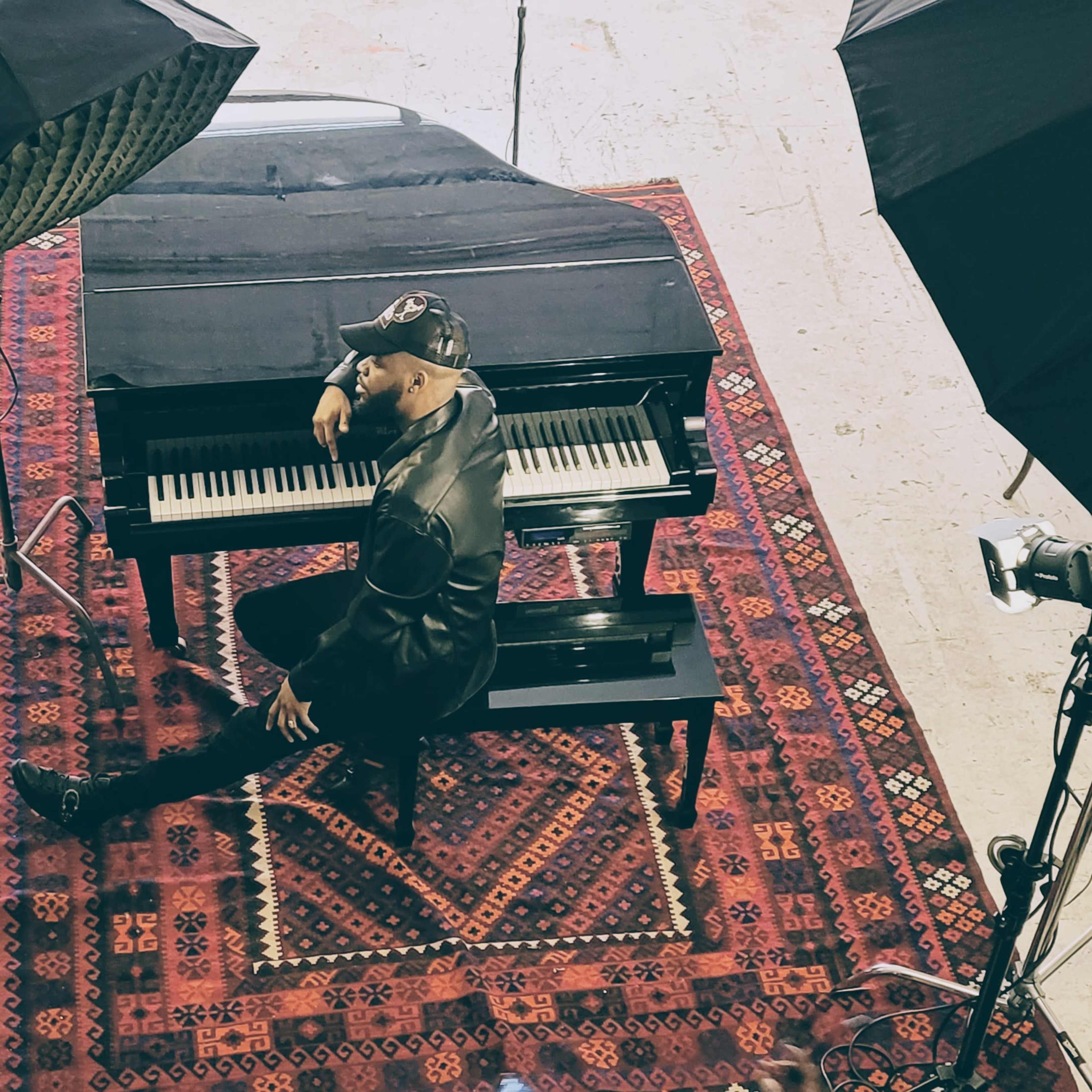 A person poses on a piano bench in front of a grand piano, surrounded by photography equipment and lighting.