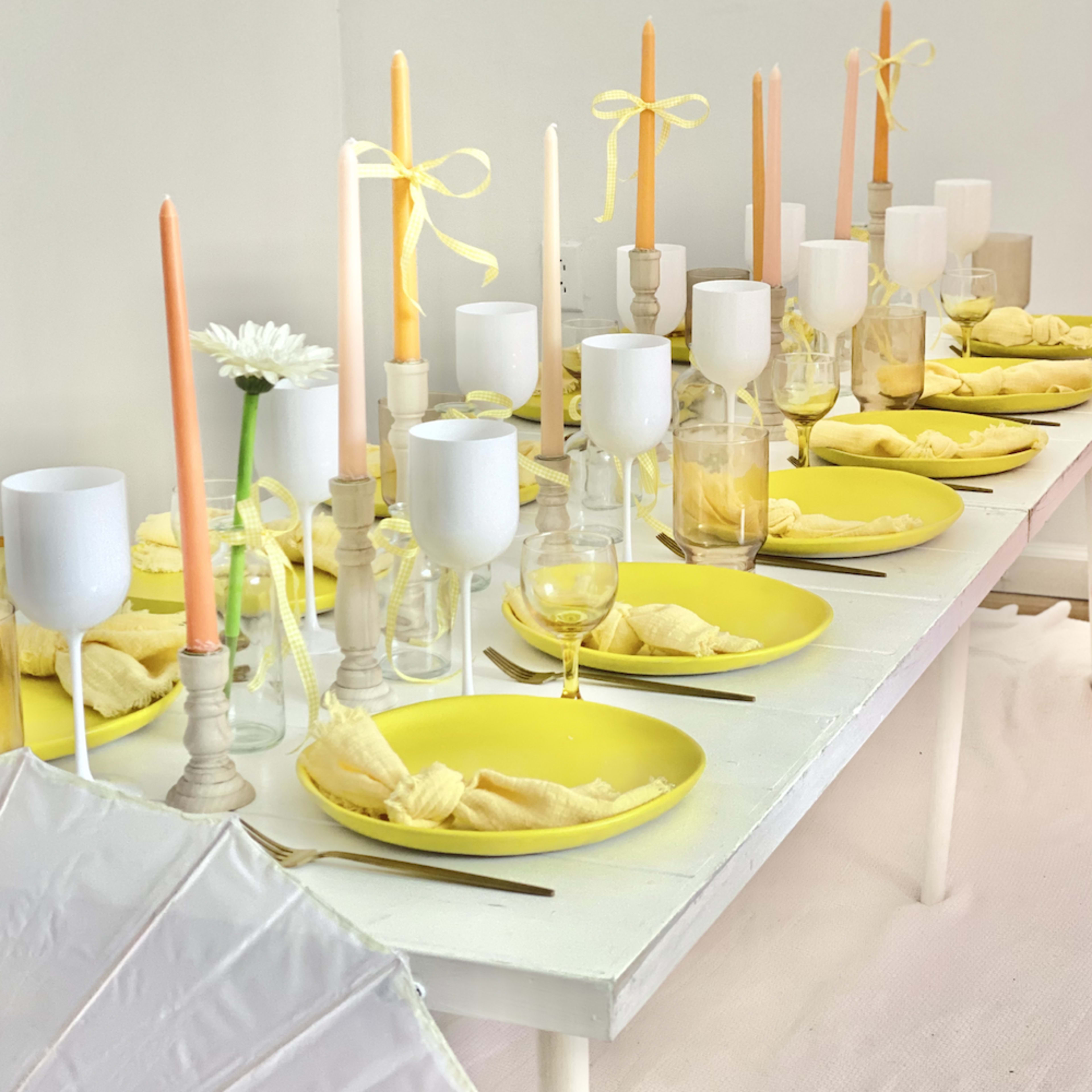 A long table is set with yellow plates, white and transparent glassware, and candles in pastel colors.