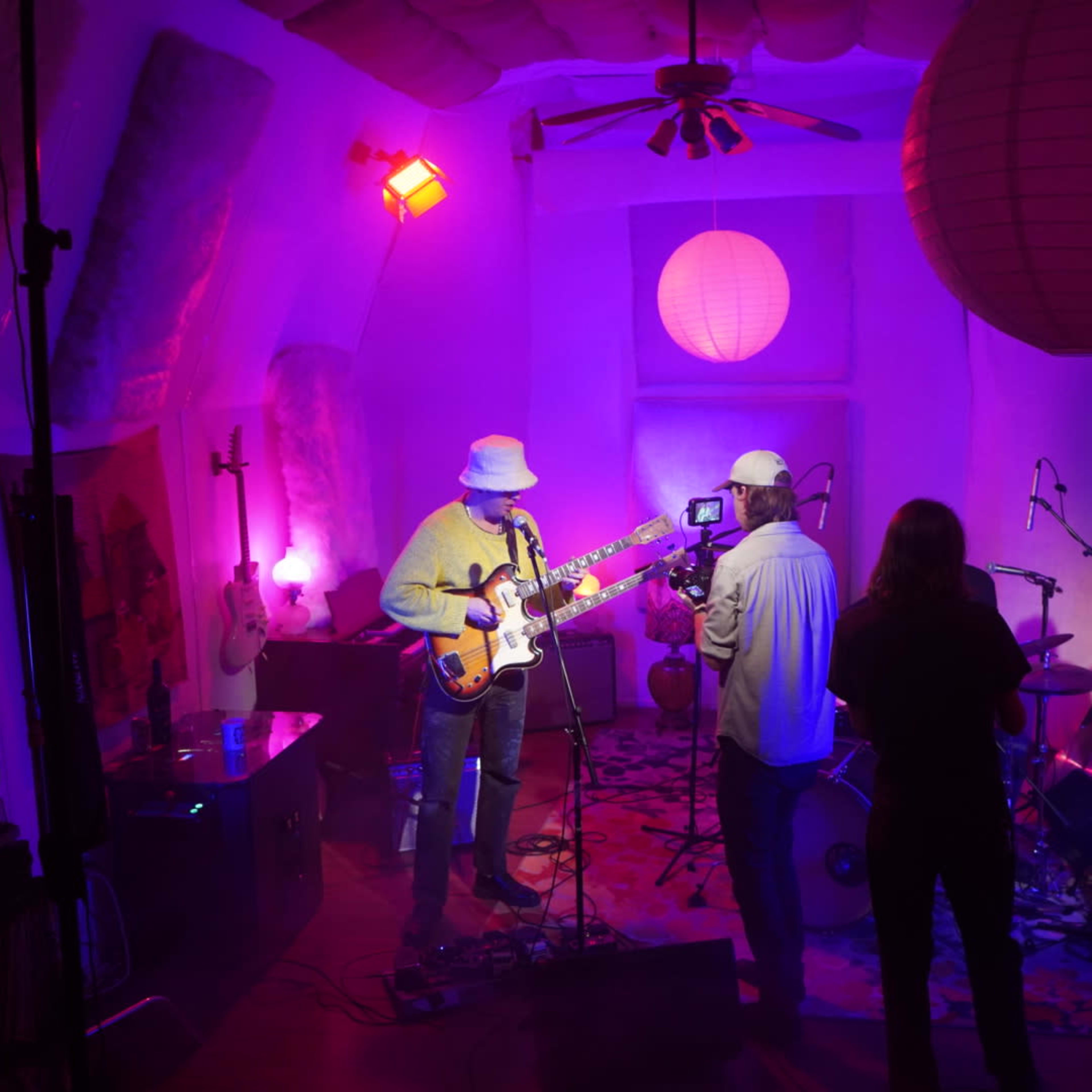 A musician performs on stage while being filmed in a colorful, dimly lit room adorned with paper lanterns and musical instruments.