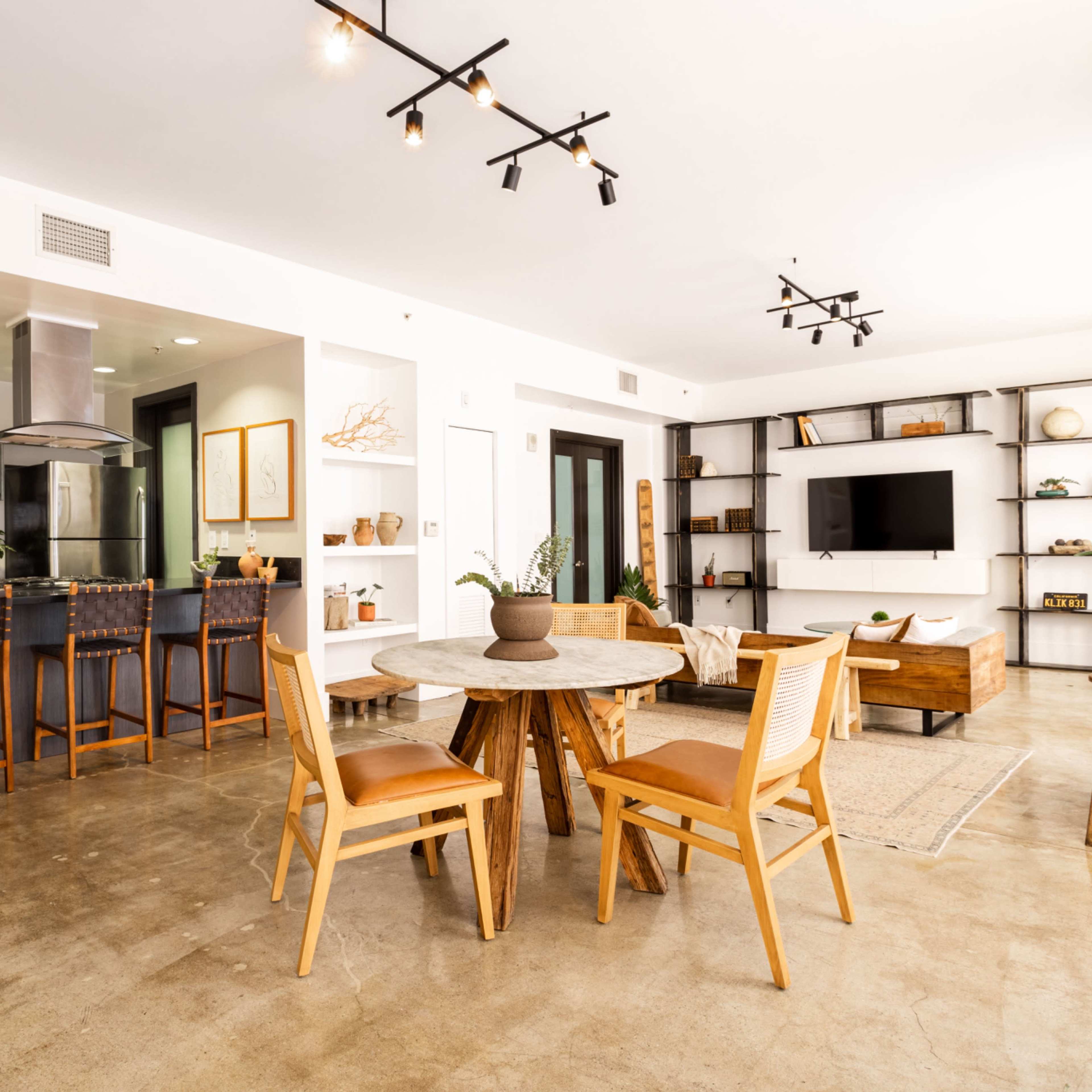 A modern open-concept living area featuring a kitchen with bar stools, a dining table, and a living space with shelves and a television.