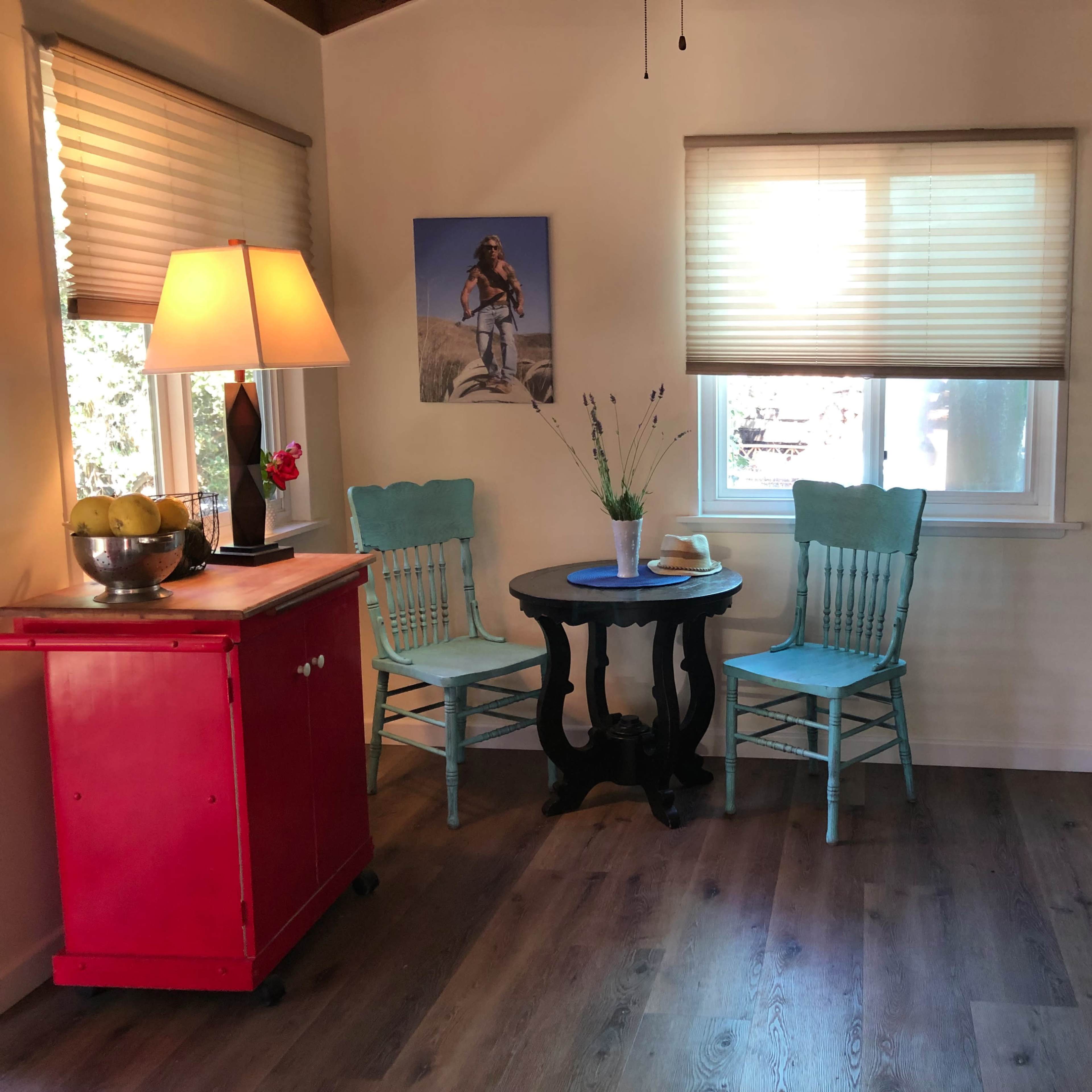 A small room features a red cabinet, a round black table with two light blue chairs, and a kitchen area with a white stove and window.