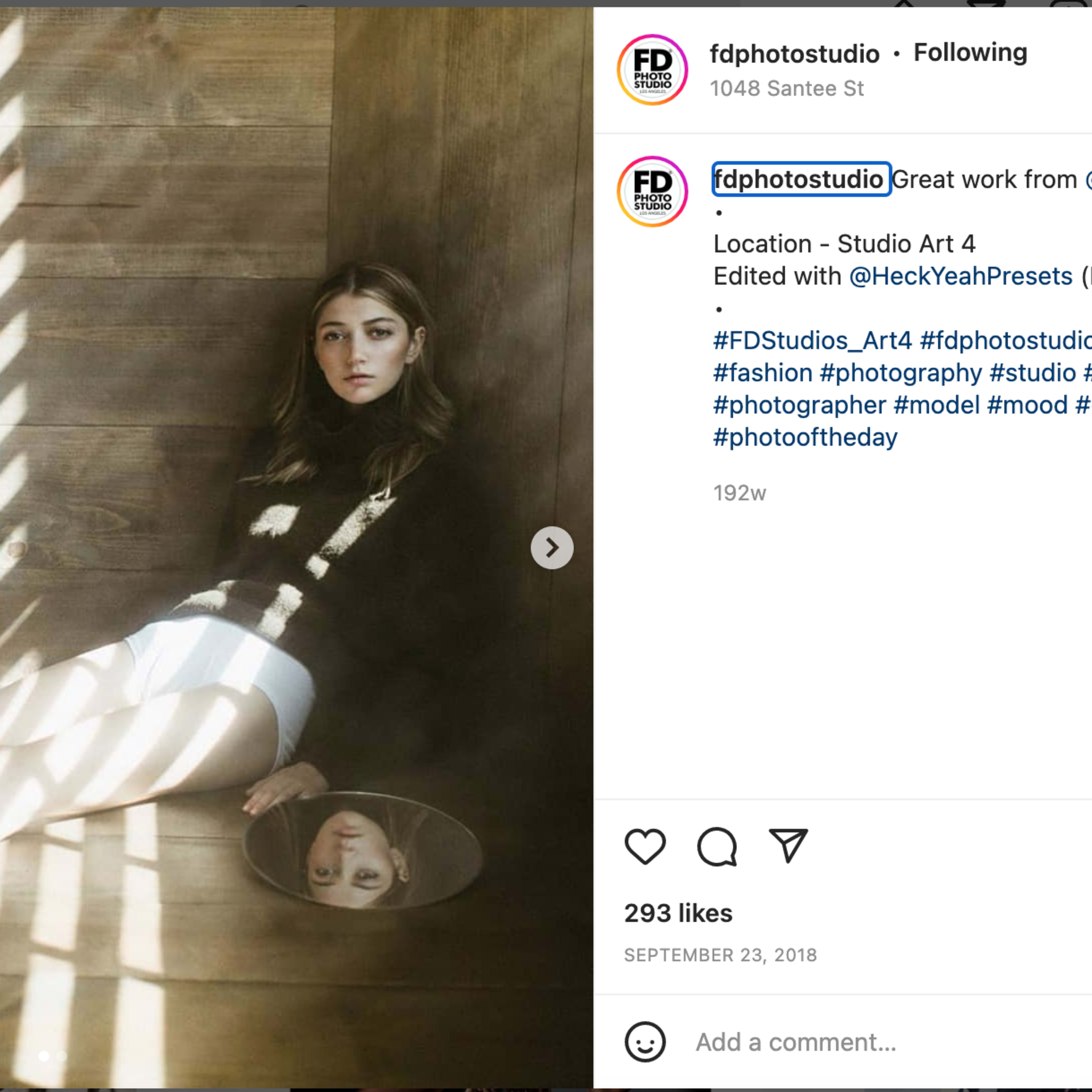 A young woman sits on the floor of a studio, gazing into a mirror while light streams through wooden slats, creating a pattern on the wall.