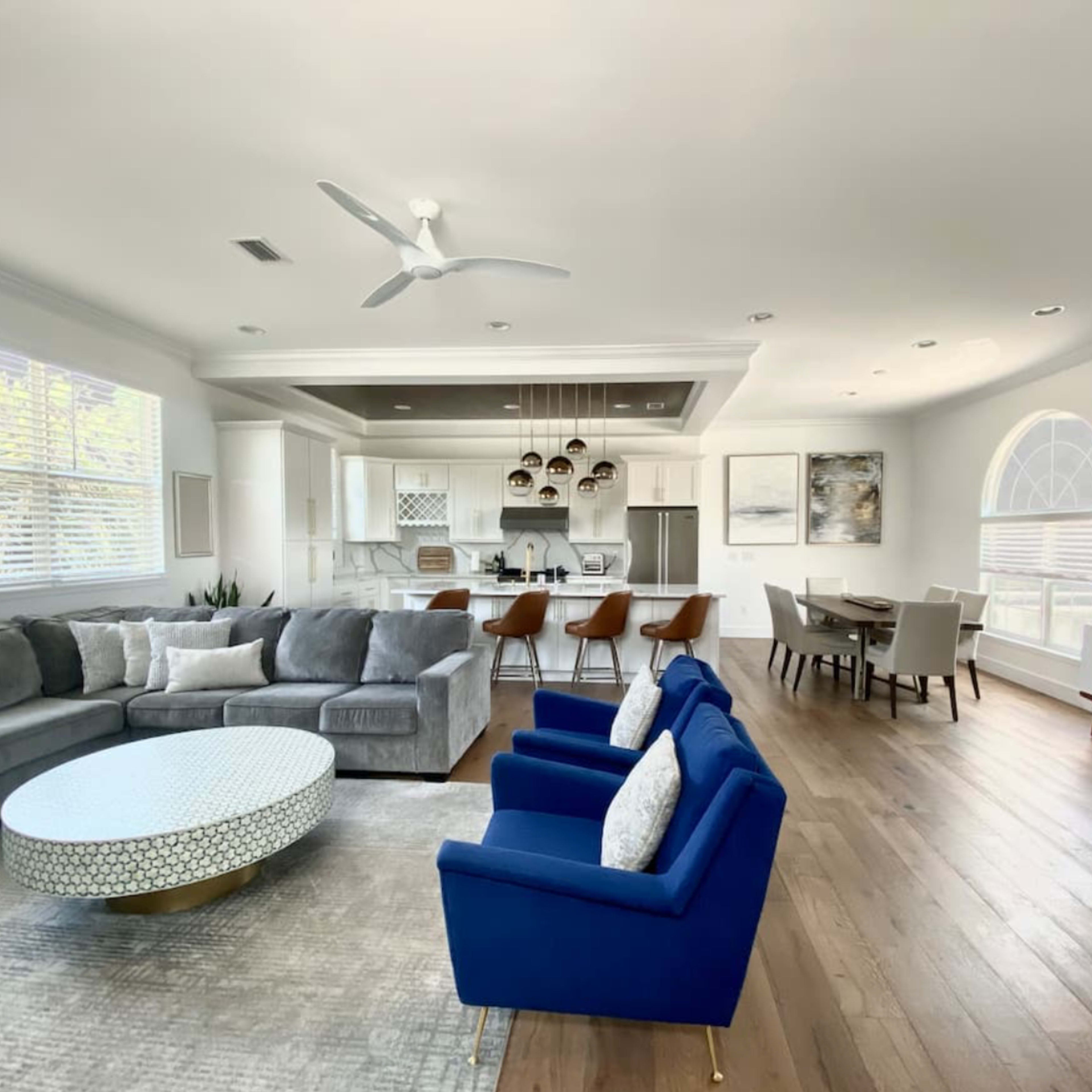 A modern living room features a sectional sofa, blue accent chairs, a circular coffee table, and an open layout connected to a dining area and kitchen.