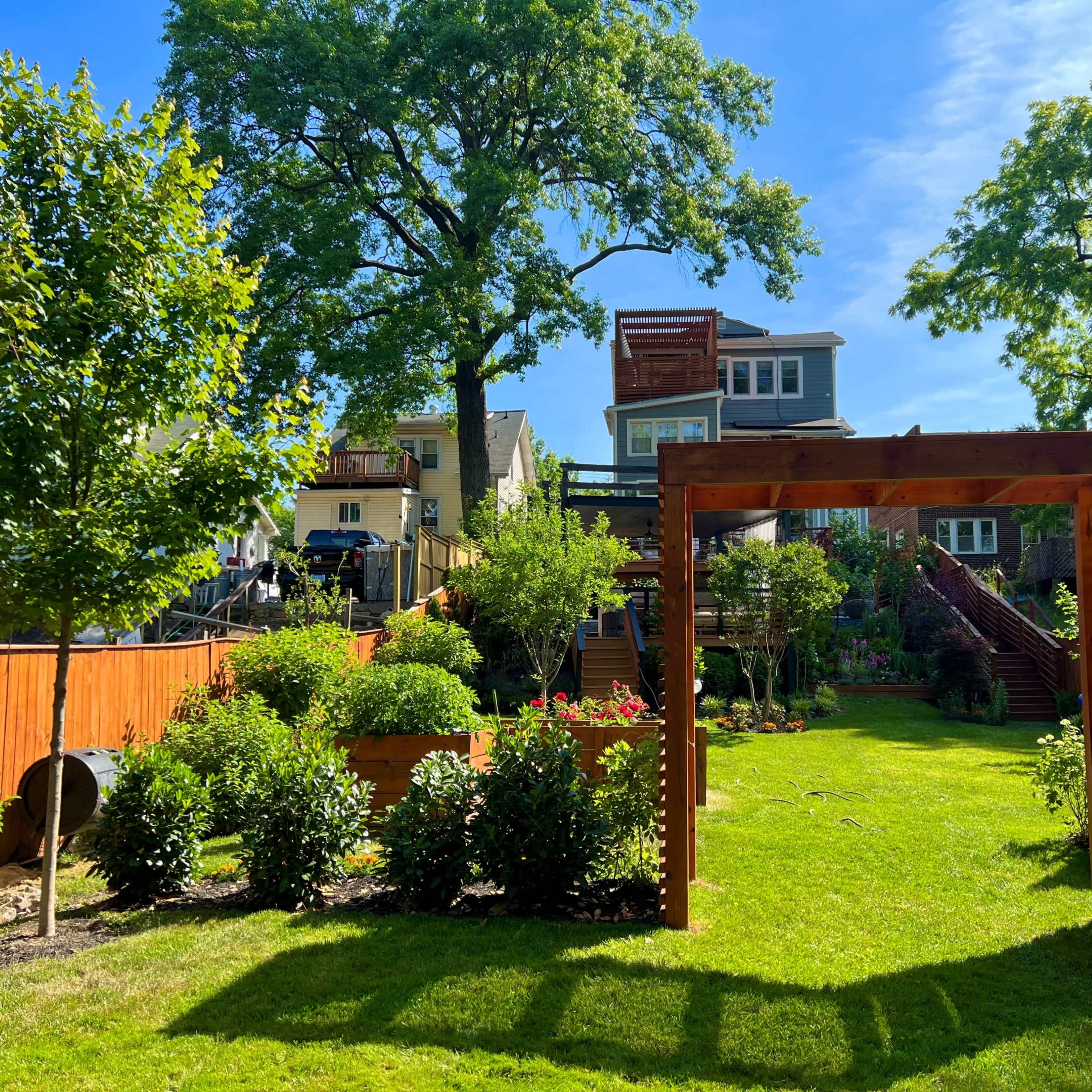 A landscaped backyard with a wooden archway leading to a garden, surrounded by trees and houses in the background.