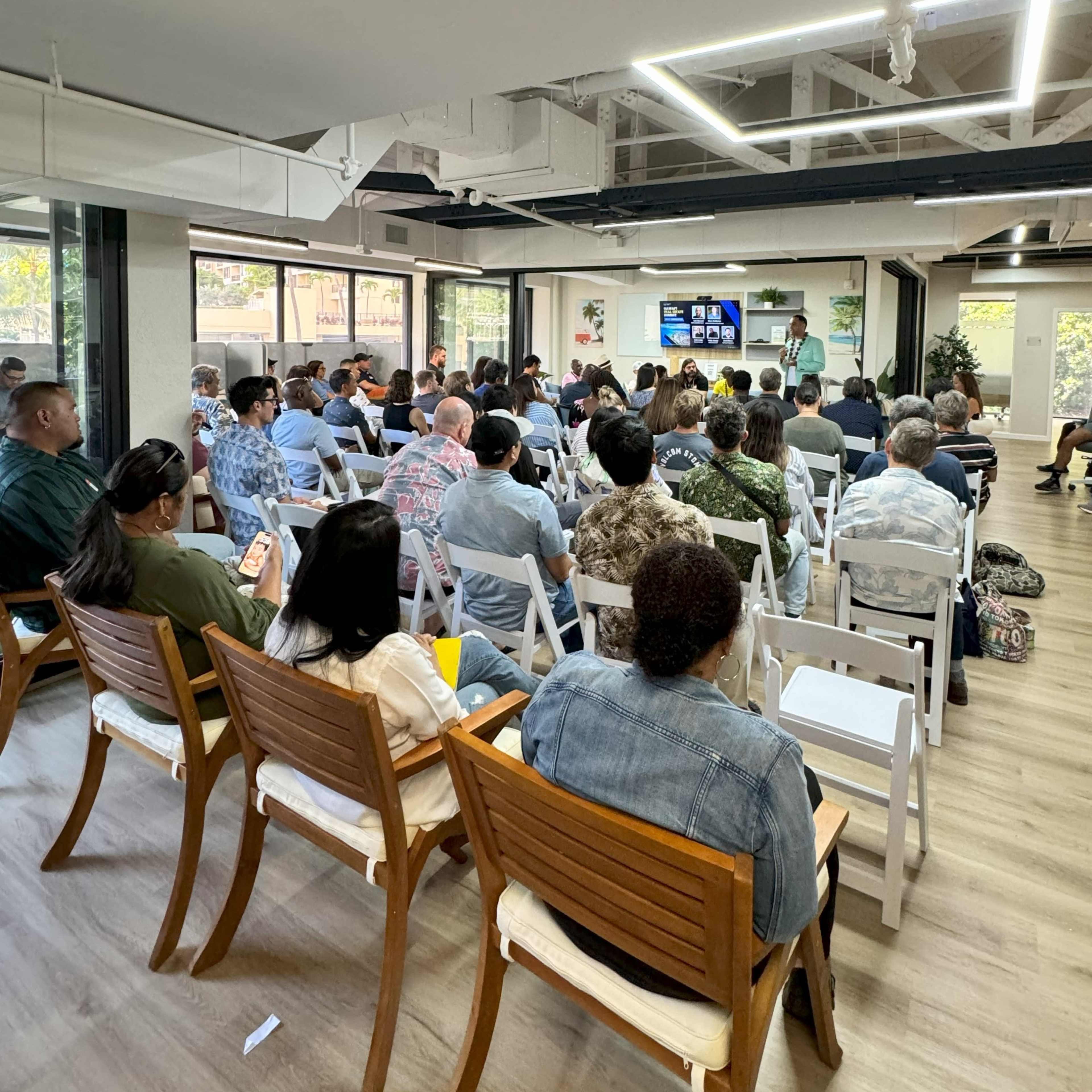 A group of attendees sits in rows of chairs facing a presenter and a screen in a modern meeting space.
